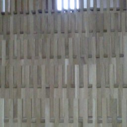 Usługi parkieciarskie - Jasne, drewniane listwy ułożone w regularny wzór, tworzące ażurową ściankę działową. Widoczne fragmenty okna w tle. Nietypowy sposób wykończenia wnętrza.