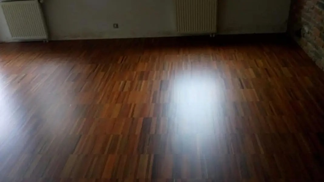 Połyskująca, ułożona pod kątem podłoga z parkietu w odcieniach brązu, z widocznymi grzejnikami przy ścianie i fragmentem ściany z cegły w prawym górnym rogu.