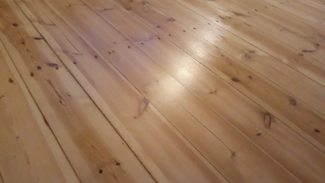 Jasne, lakierowane deski podłogowe z widocznymi słojami drewna, ułożone ukośnie. Widoczne drobne niedoskonałości i sęki, dodające naturalnego charakteru.