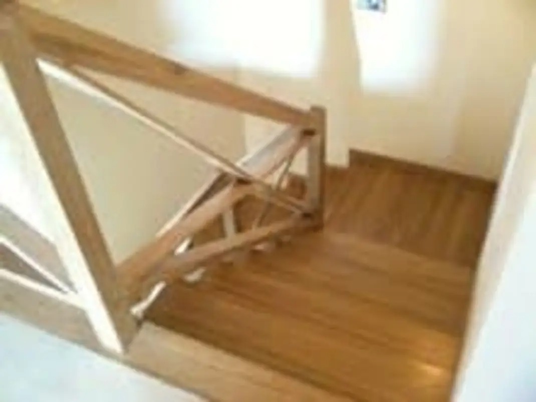 Drewniane schody z balustradą w jasnym wnętrzu. Widok z góry na stopnie i fragment ściany. Naturalne oświetlenie podkreśla strukturę drewna.