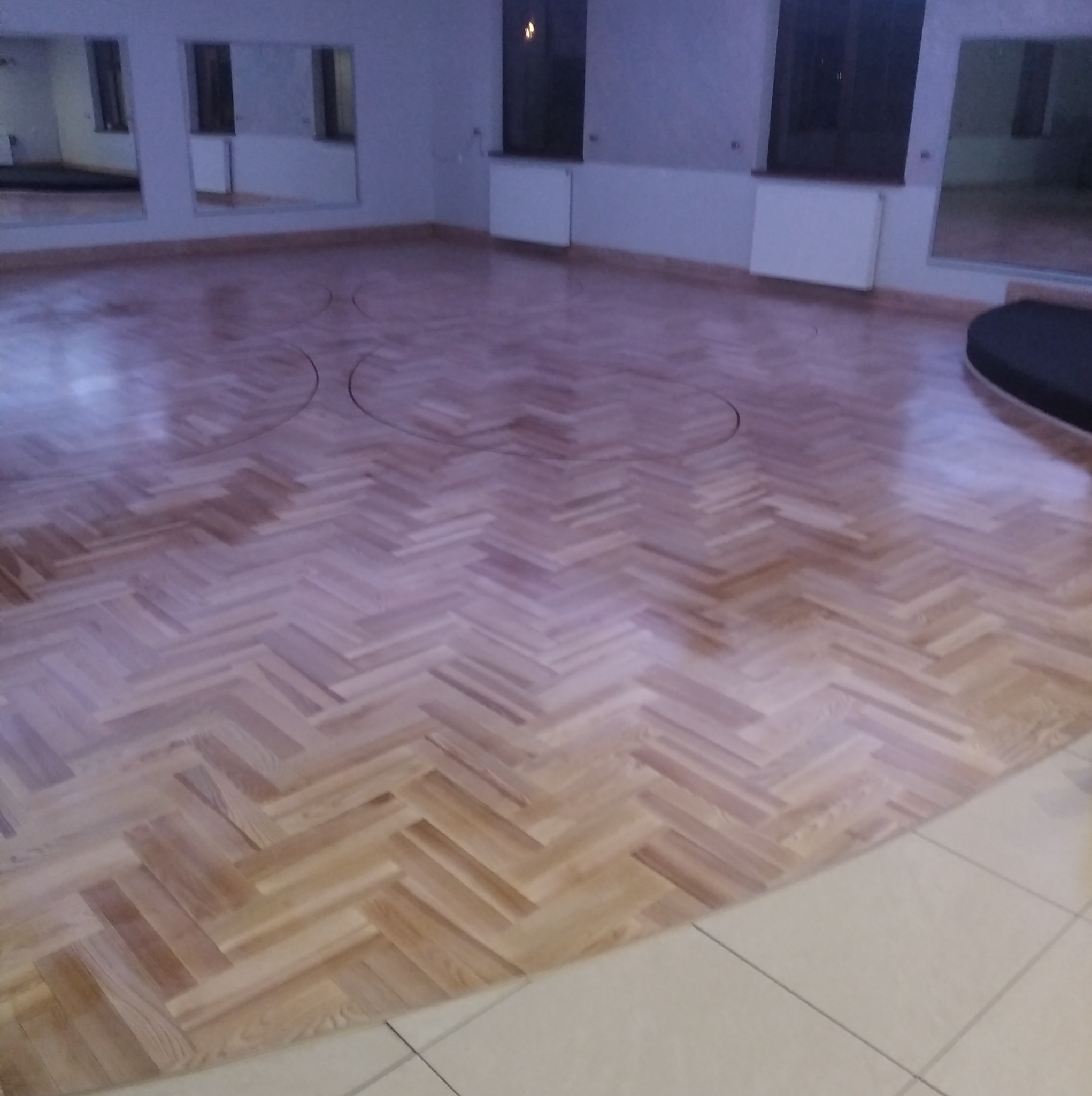 Jasny parkiet ułożony we wzór jodełki w przestronnym, jasnym pomieszczeniu z lustrami. Widoczne okrągłe ślady na podłodze i fragment białych płytek.