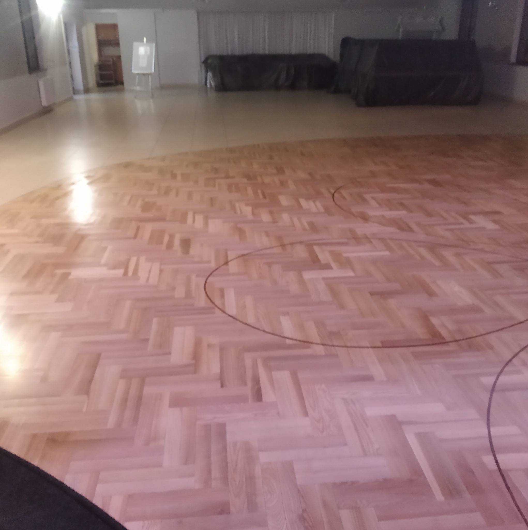 Jasny parkiet ułożony we wzór jodełki w przestronnym wnętrzu. Widoczne ciemne linie okręgów na podłodze. W tle zasłonięte okna i sprzęt.