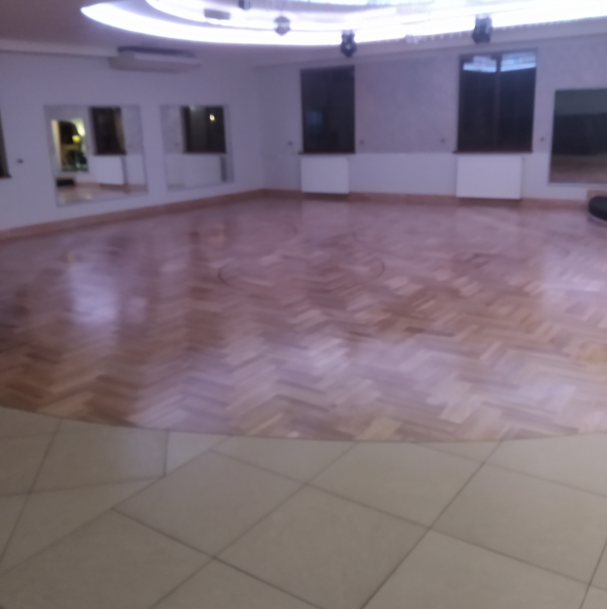 Sala balowa z parkietem ułożonym w jodełkę, lustra na ścianach i oświetlenie sufitowe. Widoczny fragment podłogi wyłożonej płytkami.