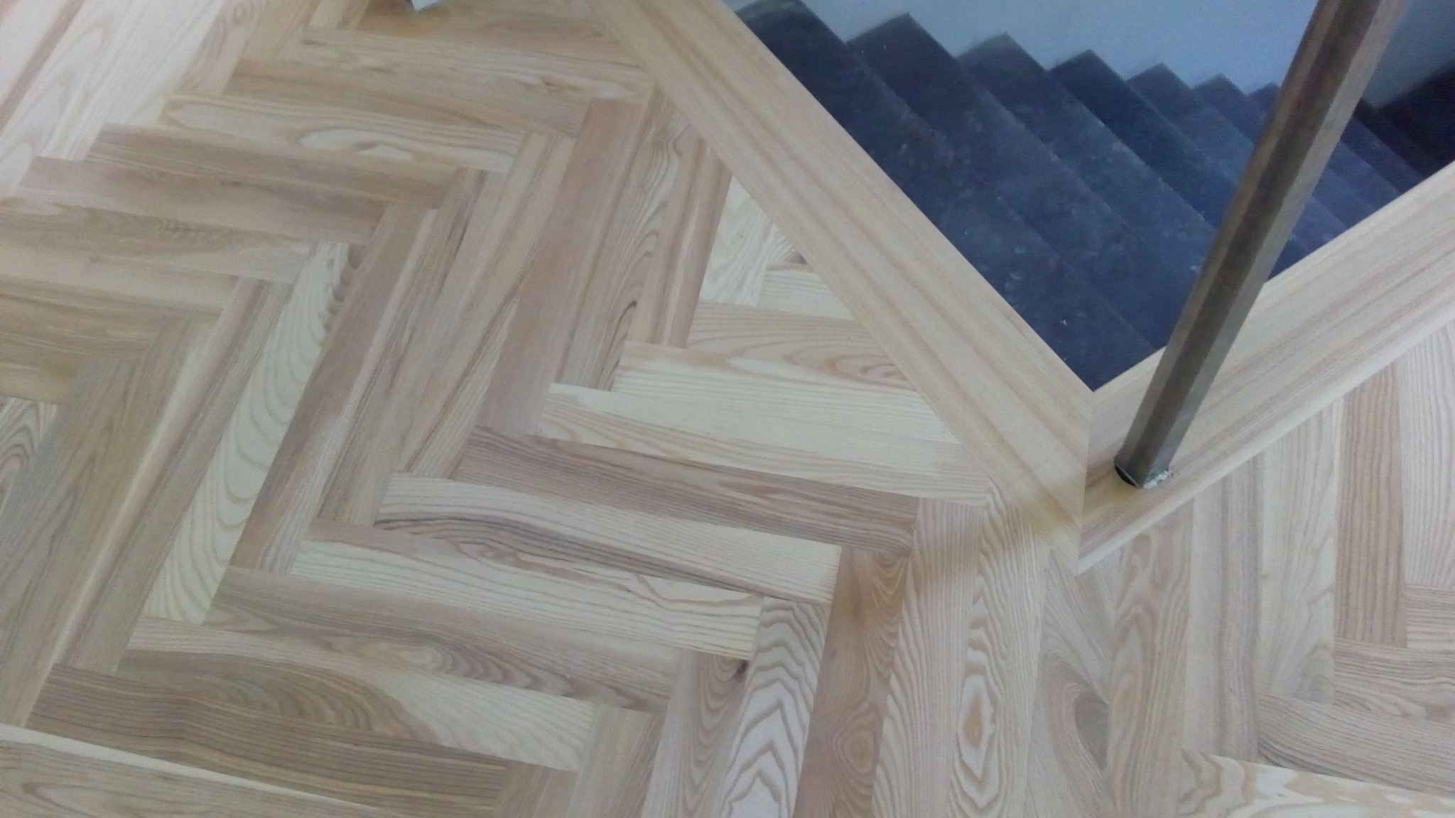 Jasny parkiet ułożony w jodełkę, z widocznym fragmentem schodów i metalowym słupkiem balustrady. Naturalne drewno z wyraźnym usłojeniem.