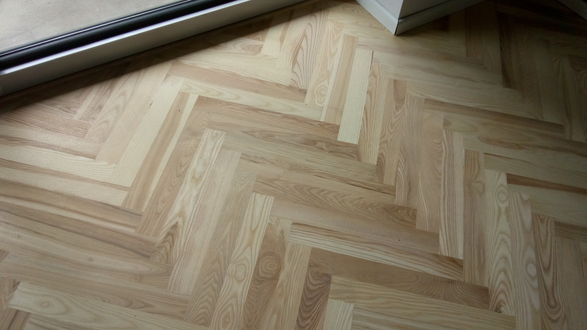 Jasny parkiet ułożony we wzór jodełki francuskiej, widoczny fragment listwy przypodłogowej i ramy okiennej. Naturalne usłojenie drewna, ciepłe oświetlenie.