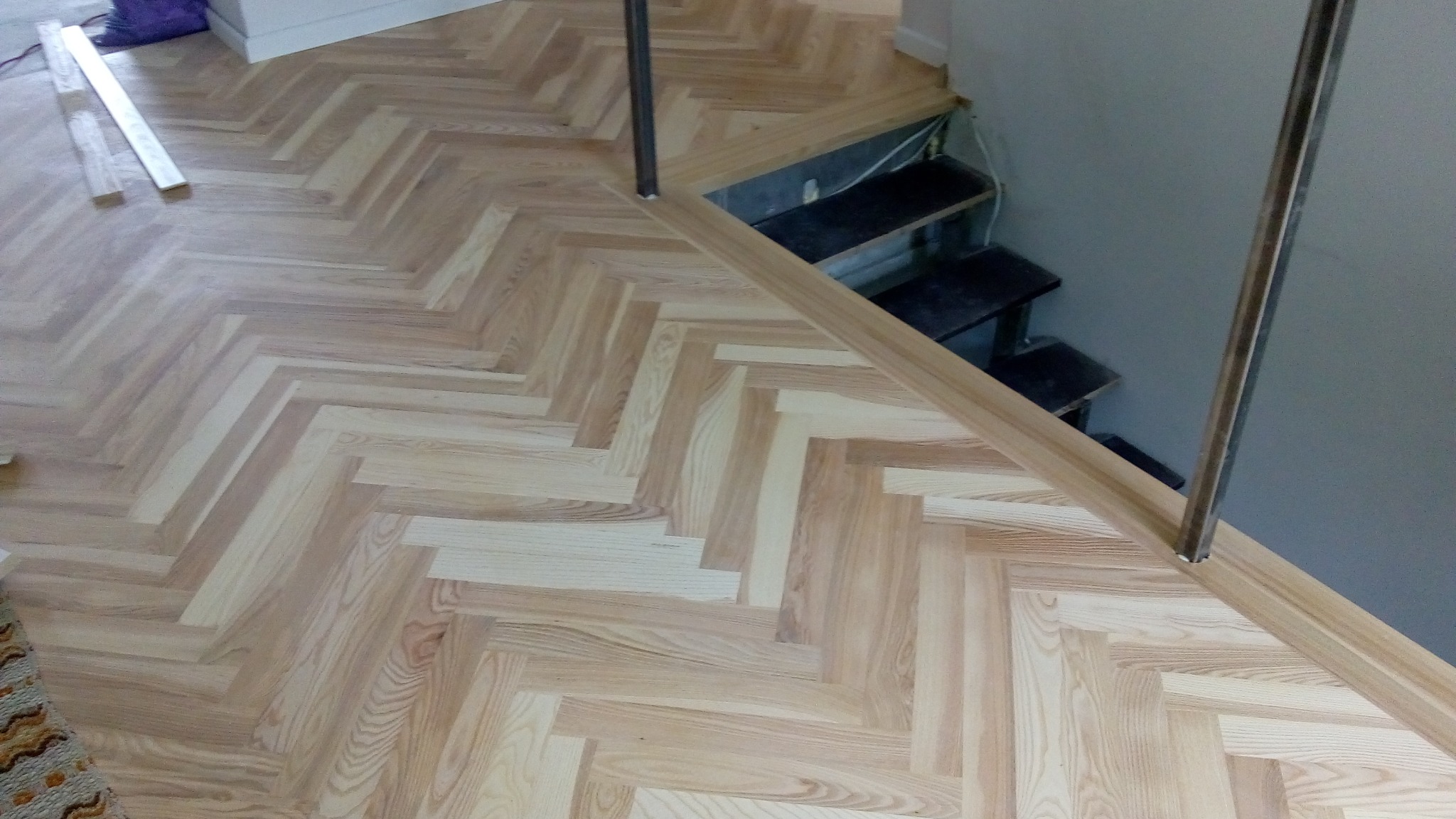 Jasny parkiet ułożony we wzór jodełki francuskiej, przechodzący w schody z metalową barierką. Widoczne deski przed montażem i fragment dywanu.