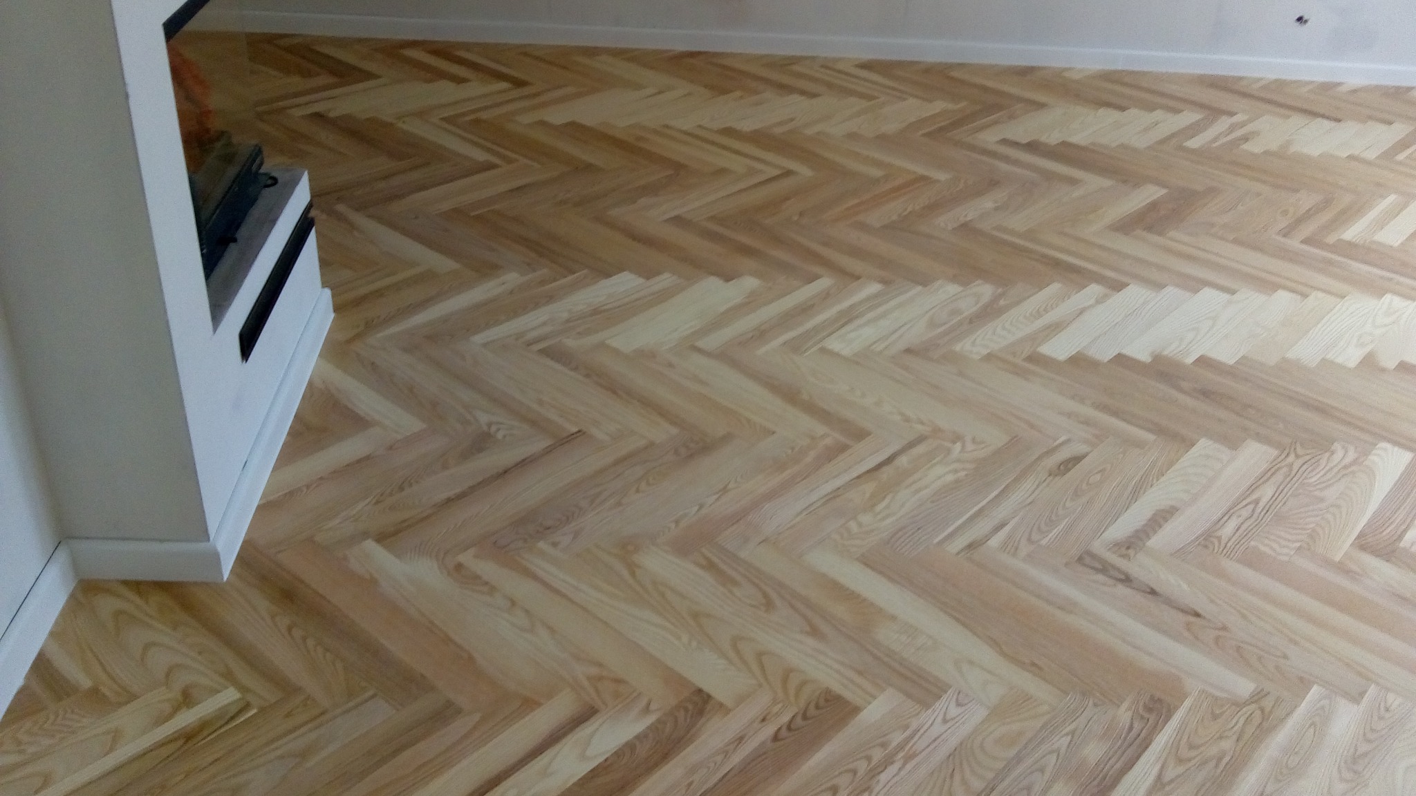 Jasny parkiet ułożony we wzór jodełki francuskiej, widoczny fragment kominka. Naturalne drewno z wyraźnym usłojeniem. Minimalistyczne wnętrze, świeżo po renowacji.