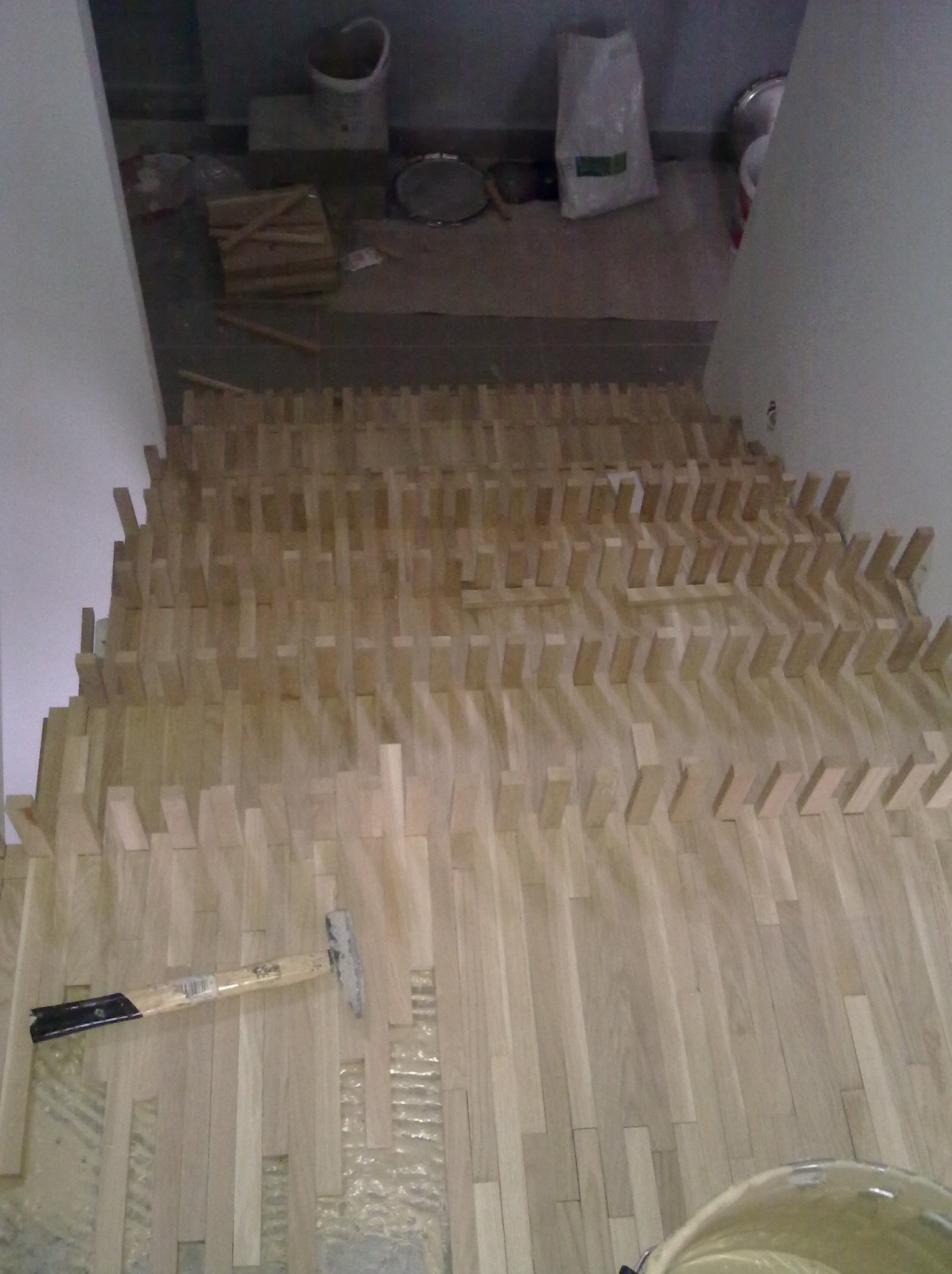 Nietypowy wzór parkietu ułożonego na podłodze, widoczny młotek i wiadro z klejem. Ujęcie z góry, ukazujące etap prac wykończeniowych w pomieszczeniu.