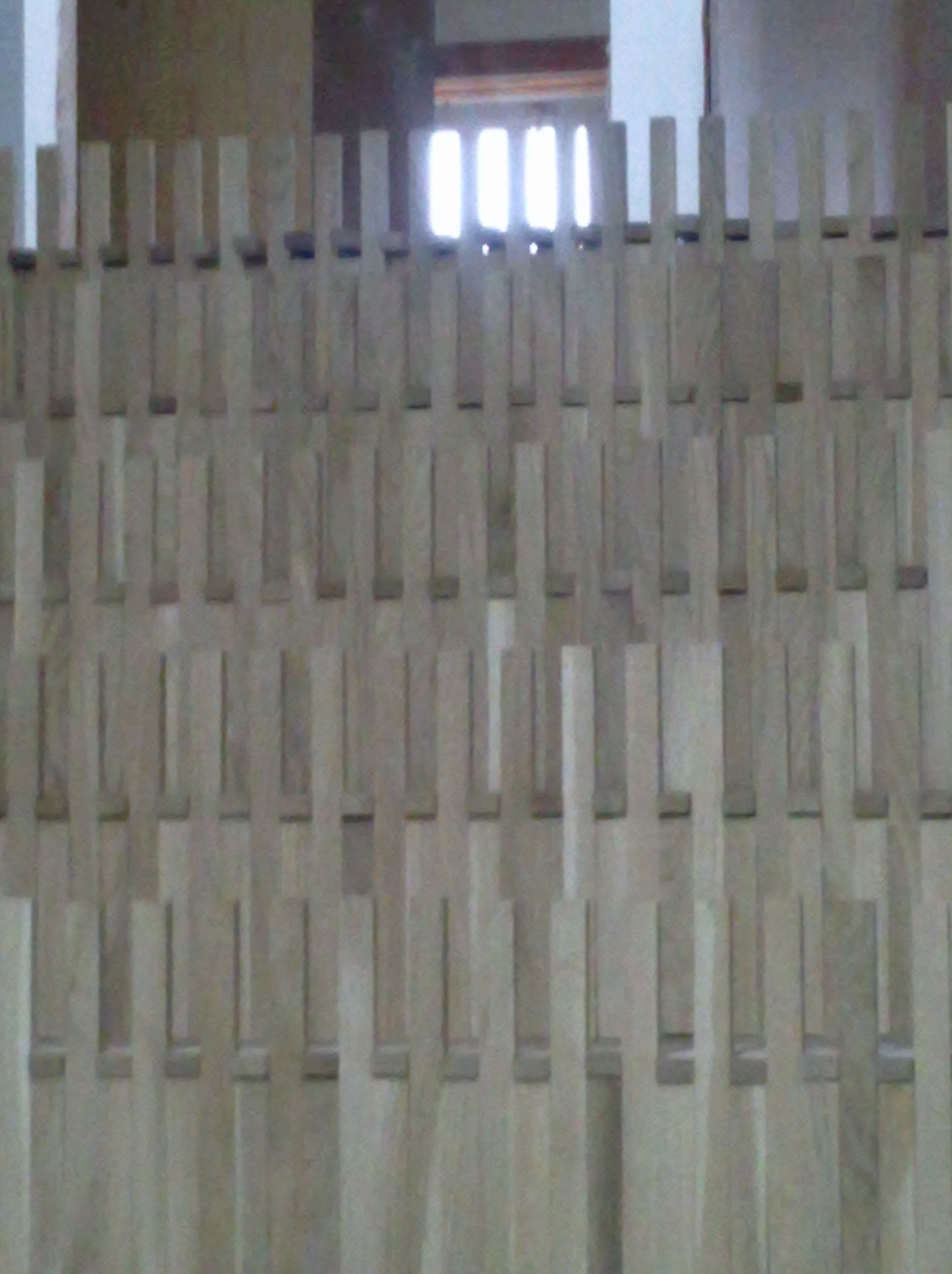 Jasne, drewniane listwy ułożone w regularny wzór, tworzące ażurową ściankę działową. Widoczne fragmenty okna w tle. Nietypowy sposób wykończenia wnętrza.