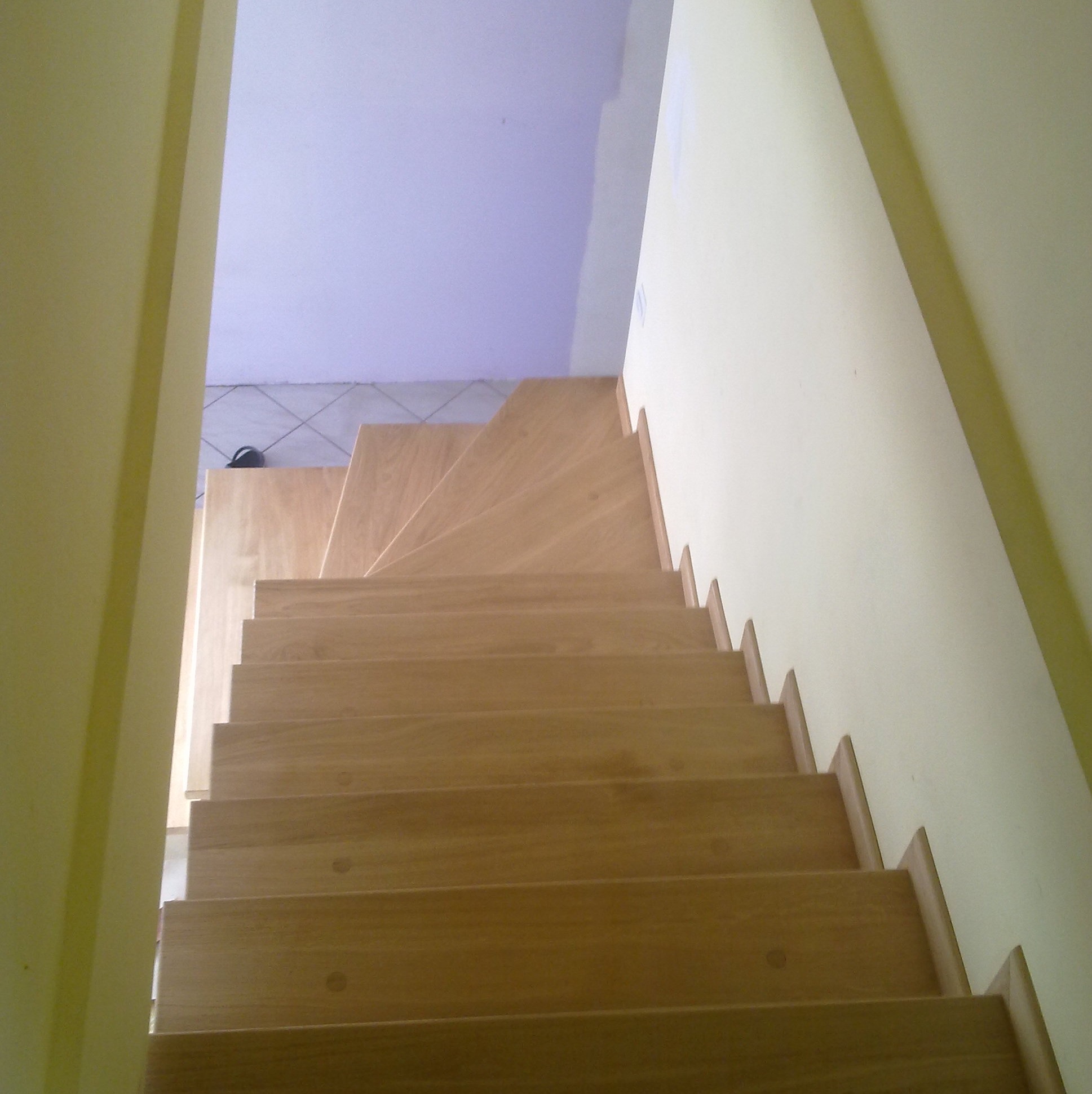 Jasne, drewniane schody z widokiem z góry, prowadzące w górę. Widoczne detale wykończenia i kąt ujęcia podkreślający perspektywę. Ściany w odcieniach żółci i fioletu.