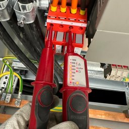 4MP SPÓŁKA Z OGRANICZONĄ ODPOWIEDZIALNOŚCIĄ - Pomiar instalacji elektrycznej w Gdyni. Tester Benning w rękawicy dotyka czerwonych złączek L1, L2, L3 i N. Widoczne kable i elementy instalacji.