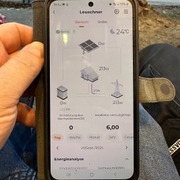 4MP SPÓŁKA Z OGRANICZONĄ ODPOWIEDZIALNOŚCIĄ - Ekran smartfona prezentuje analizę energii z instalacji fotowoltaicznej, z widocznymi danymi o produkcji, zużyciu i mocy. W tle fragment otoczenia zewnętrznego.