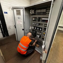 4MP SPÓŁKA Z OGRANICZONĄ ODPOWIEDZIALNOŚCIĄ - Elektryk w kamizelce BHP kuca przed otwartą szafą sterowniczą, wykonując prace instalacyjne. Widoczne liczne przewody i elementy elektryczne wewnątrz szafy.