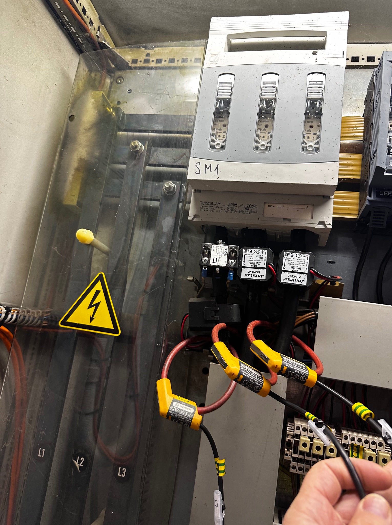 Szafa elektryczna z bezpiecznikami, okablowaniem i żółtym znakiem ostrzegawczym wysokiego napięcia. Widoczne elementy pomiarowe i oznaczenia L1, L2, L3.