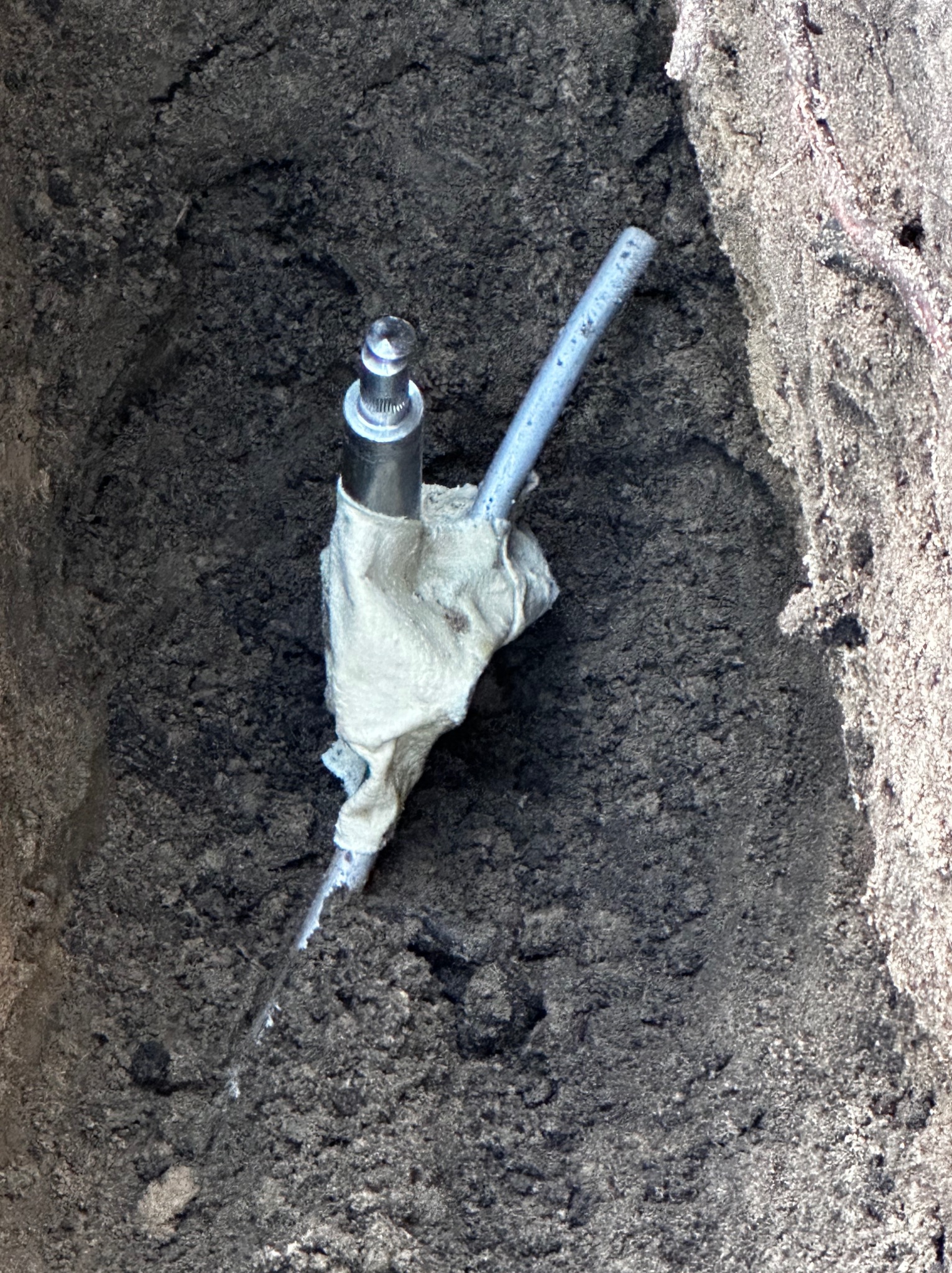 Uziemienie instalacji odgromowej w wykopie, zabezpieczone materiałem izolacyjnym. Widoczny fragment przewodu i złącze. Tło stanowi ziemia.