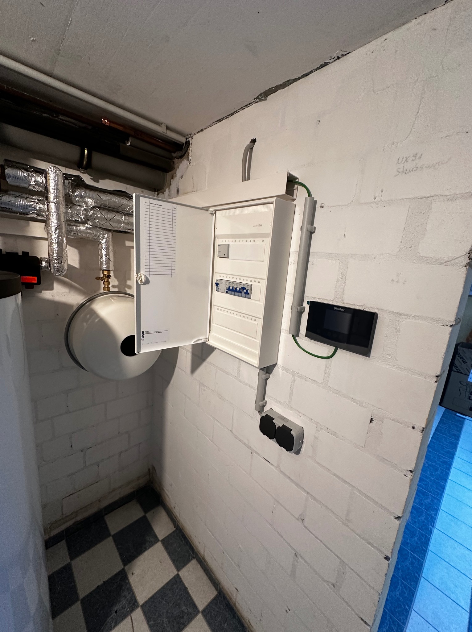 Otwarta skrzynka elektryczna Hager na białej ścianie z cegły, obok czarny panel sterowania Vaillant. Widoczne rury i instalacje. Podłoga w szachownicę.