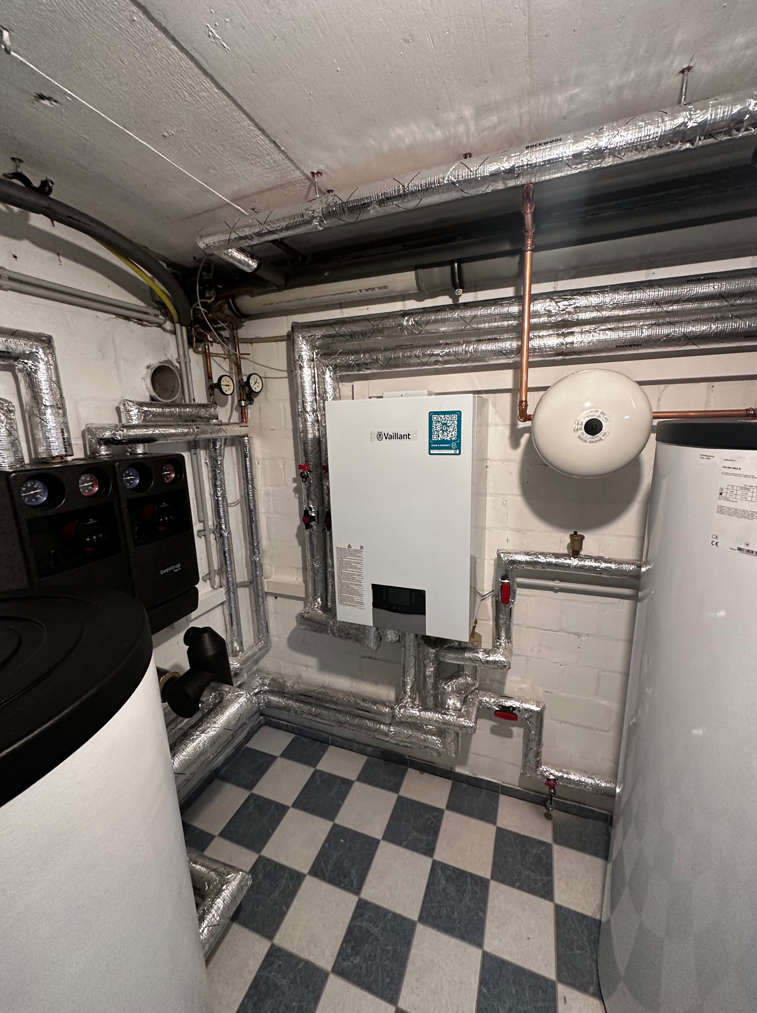 Kotłownia z nowoczesnym piecem Vaillant, zbiornikiem buforowym i instalacją grzewczą z rur w izolacji, na tle białej ściany z cegły i szachownicy na podłodze.