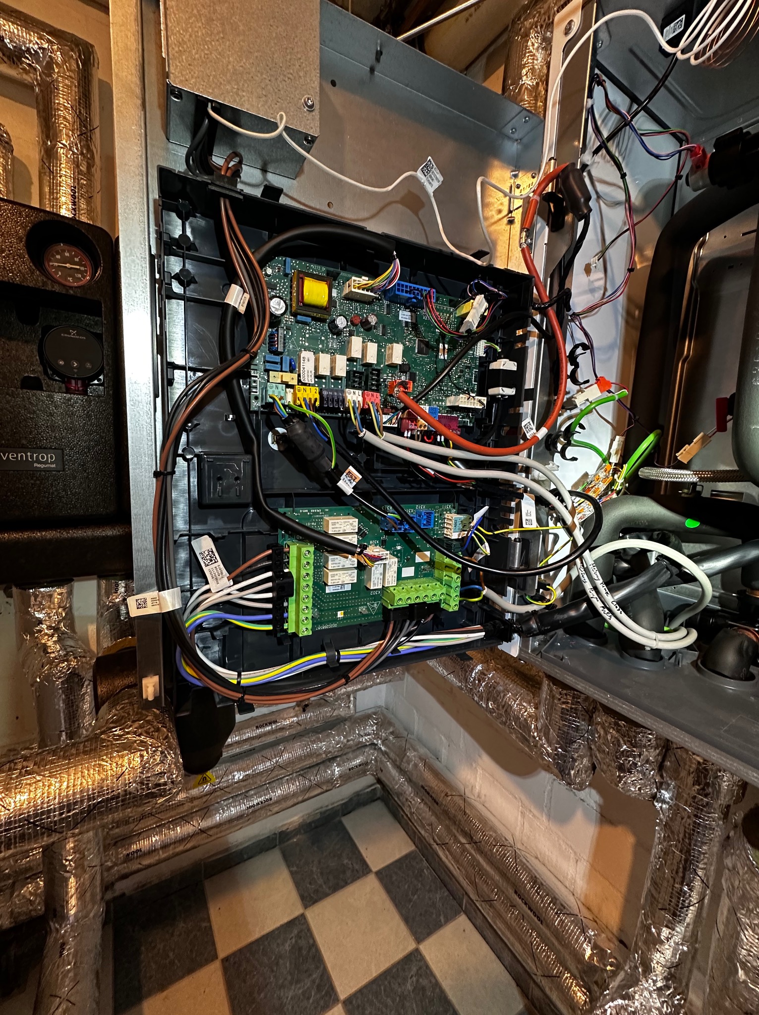 Otwarta skrzynka sterownicza z widocznymi płytkami drukowanymi, kablami i przewodami. W tle izolowane rury i fragment podłogi w szachownicę.