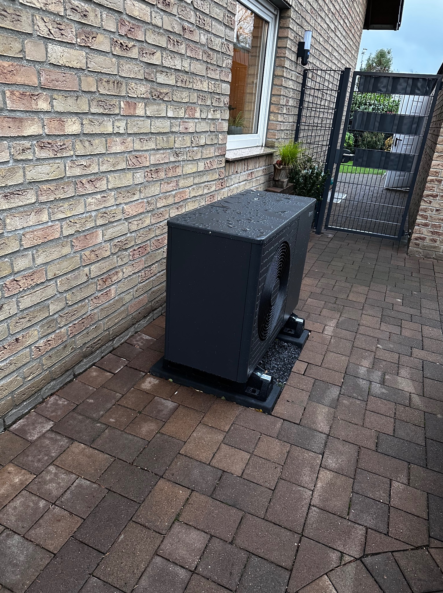 Czarna pompa ciepła na zewnątrz budynku z cegły, widoczna brama w tle. Urządzenie stoi na czarnych podstawach z drobnym żwirem. Widoczne krople deszczu na obudowie.