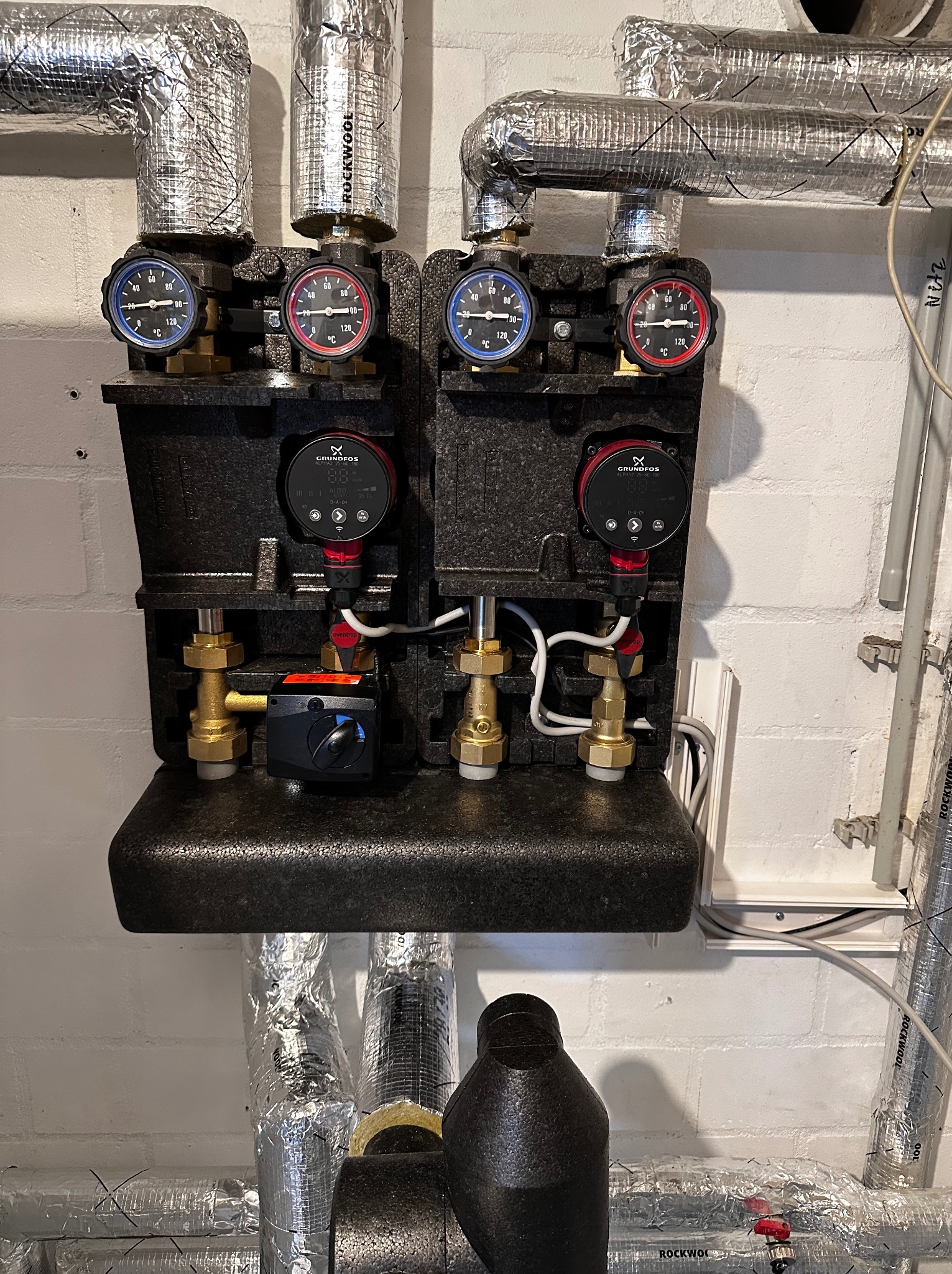 Złożona instalacja grzewcza z czarną izolacją, manometrami i pompami obiegowymi Grundfos, rury w srebrnej izolacji Rockwool na tle białej ściany z cegły.