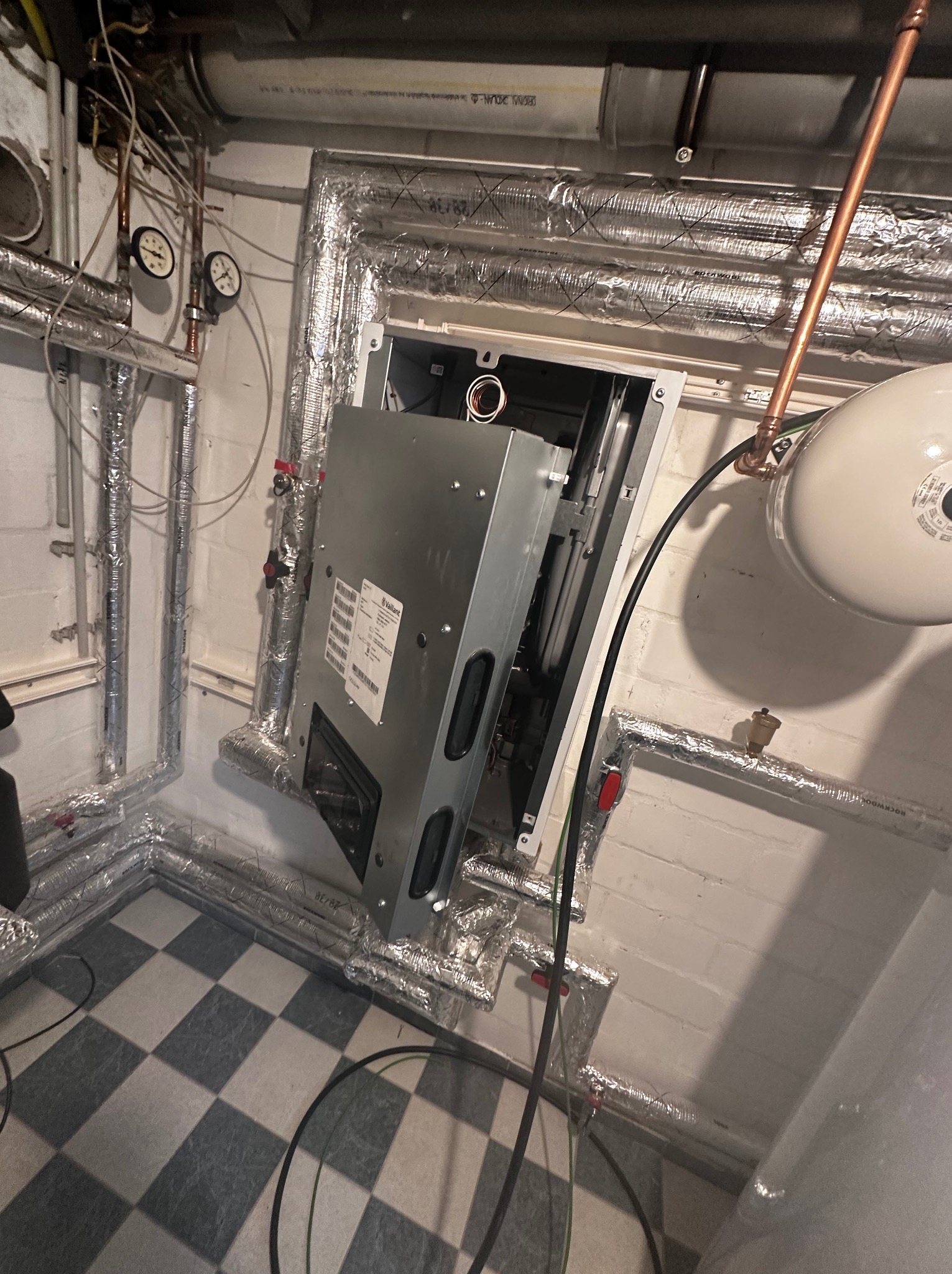 Otwarta obudowa urządzenia Vaillant w kotłowni z izolowanymi rurami, manometrami i zbiornikiem wyrównawczym. Widoczna szachownica na podłodze i fragment instalacji.