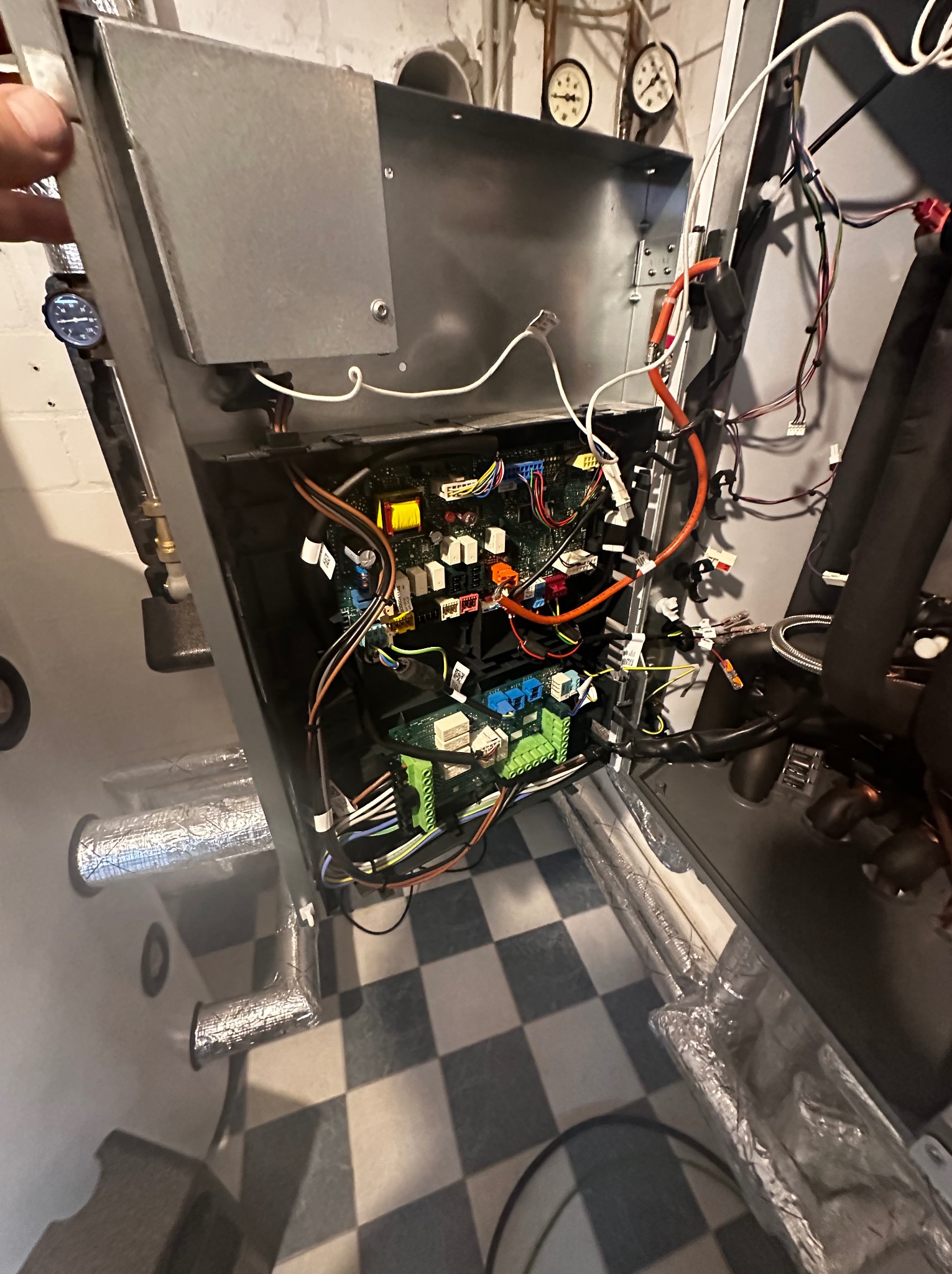 Otwarta obudowa urządzenia z widoczną płytą sterującą, liczne przewody i złącza. W tle ściana z manometrami i rury w izolacji. Podłoga w szachownicę.