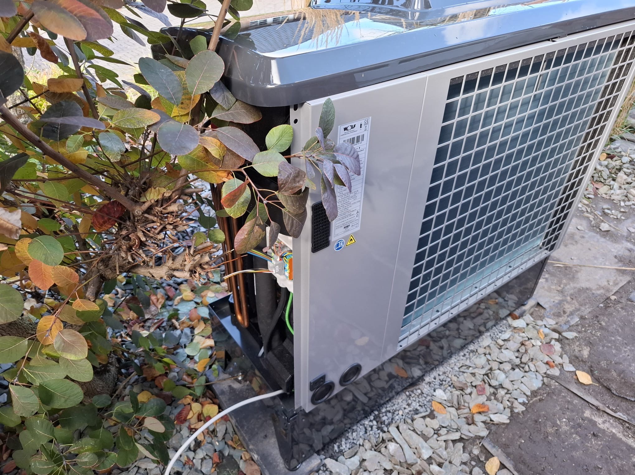 Szara pompa ciepła typu monoblok, widoczna z boku, z odsłoniętymi elementami instalacji, na tle kamieni i roślinności. Widoczna tabliczka znamionowa.