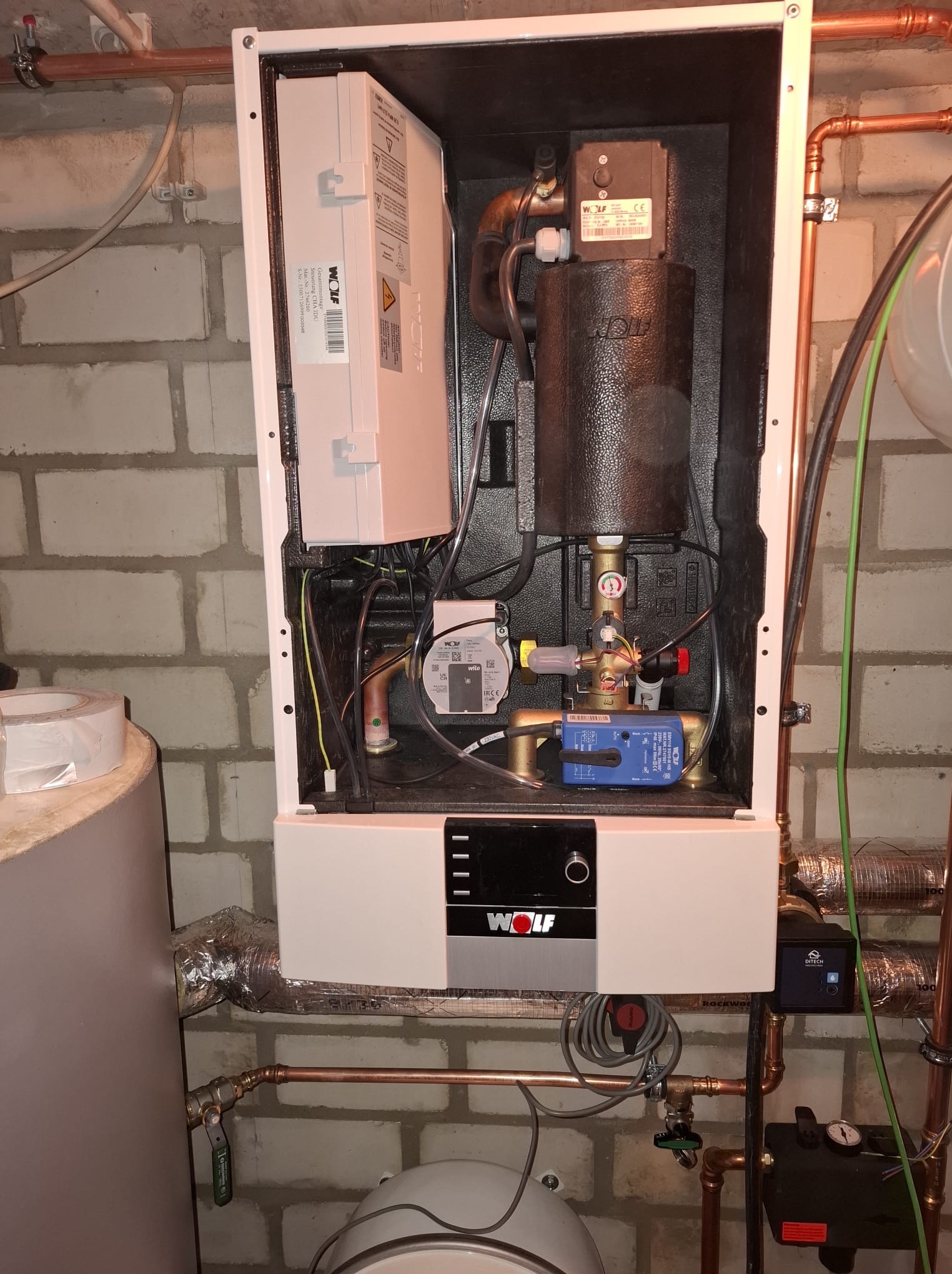 Zamknięta obudowa kotła grzewczego Wolf z widocznymi elementami instalacji, rurami miedzianymi i przewodami. Tło stanowi ściana z cegły.