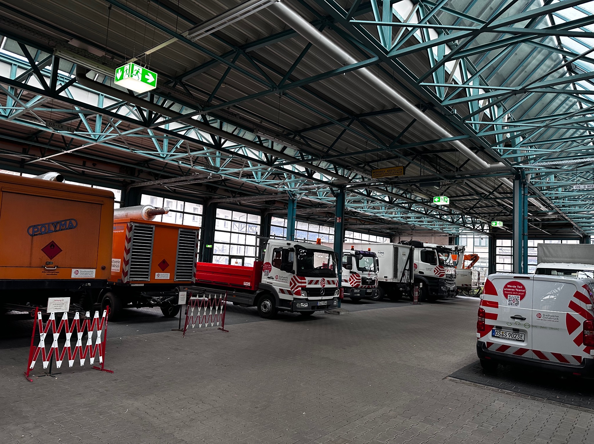 Wnętrze hali z pojazdami komunalnymi, widoczne oznakowania i oświetlenie awaryjne pod sufitem. Konstrukcja stalowa dachu z widocznymi przewodami.