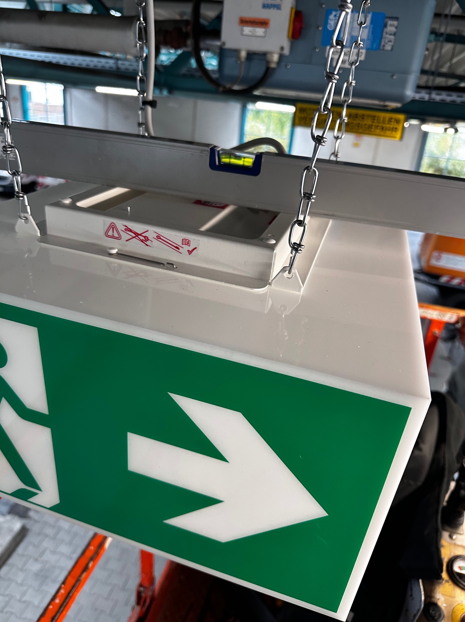 Zawieszony na łańcuchach, podświetlany znak ewakuacyjny z zieloną strzałką i piktogramem osoby biegnącej, zamontowany pod sufitem. Widoczna poziomica i elementy instalacji.