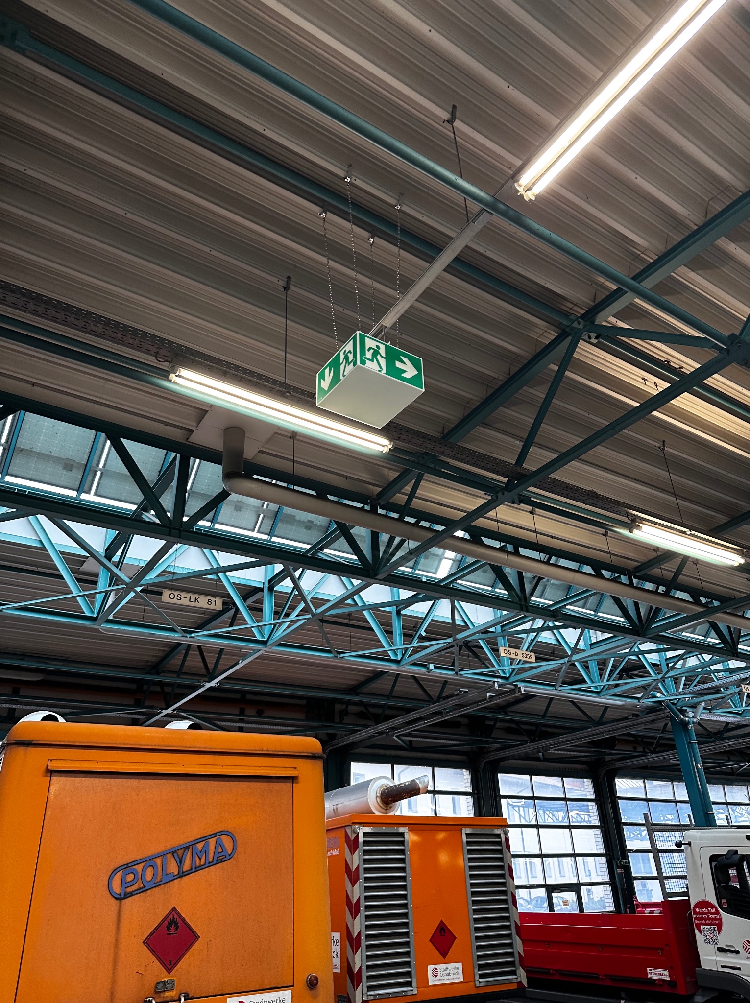 Oświetlenie hali przemysłowej z lampami jarzeniowymi, znakiem ewakuacyjnym i konstrukcją stalową pod dachem. Widoczne pomarańczowe agregaty i okna w tle.