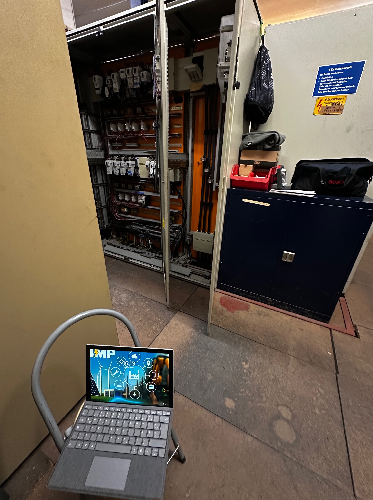 Otwarta szafa elektryczna z widocznymi przewodami i elementami sterującymi, laptop na krześle z logo firmy na ekranie, narzędzia i torba obok szafy.