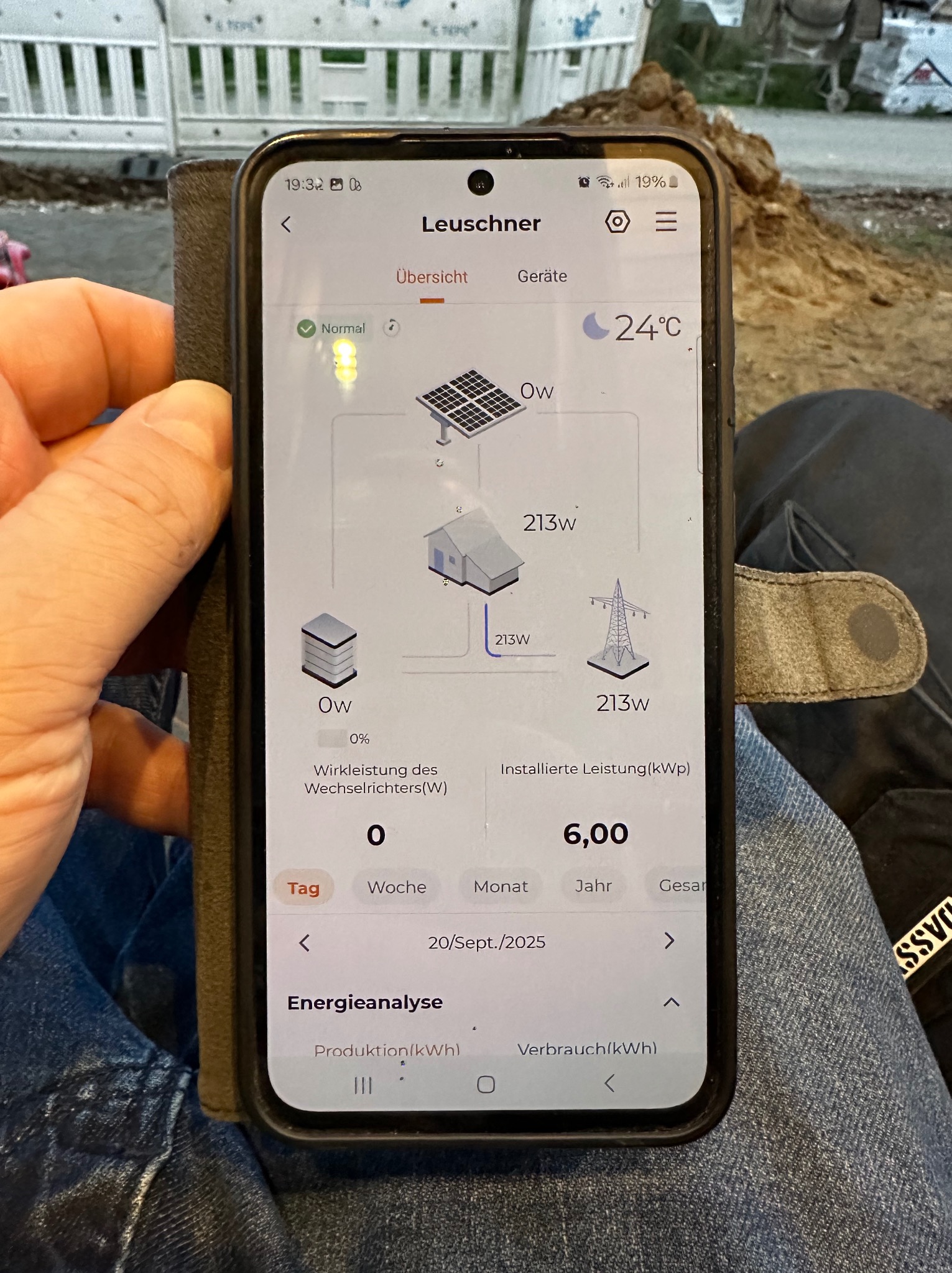 Ekran smartfona prezentuje analizę energii z instalacji fotowoltaicznej, z widocznymi danymi o produkcji, zużyciu i mocy. W tle fragment otoczenia zewnętrznego.