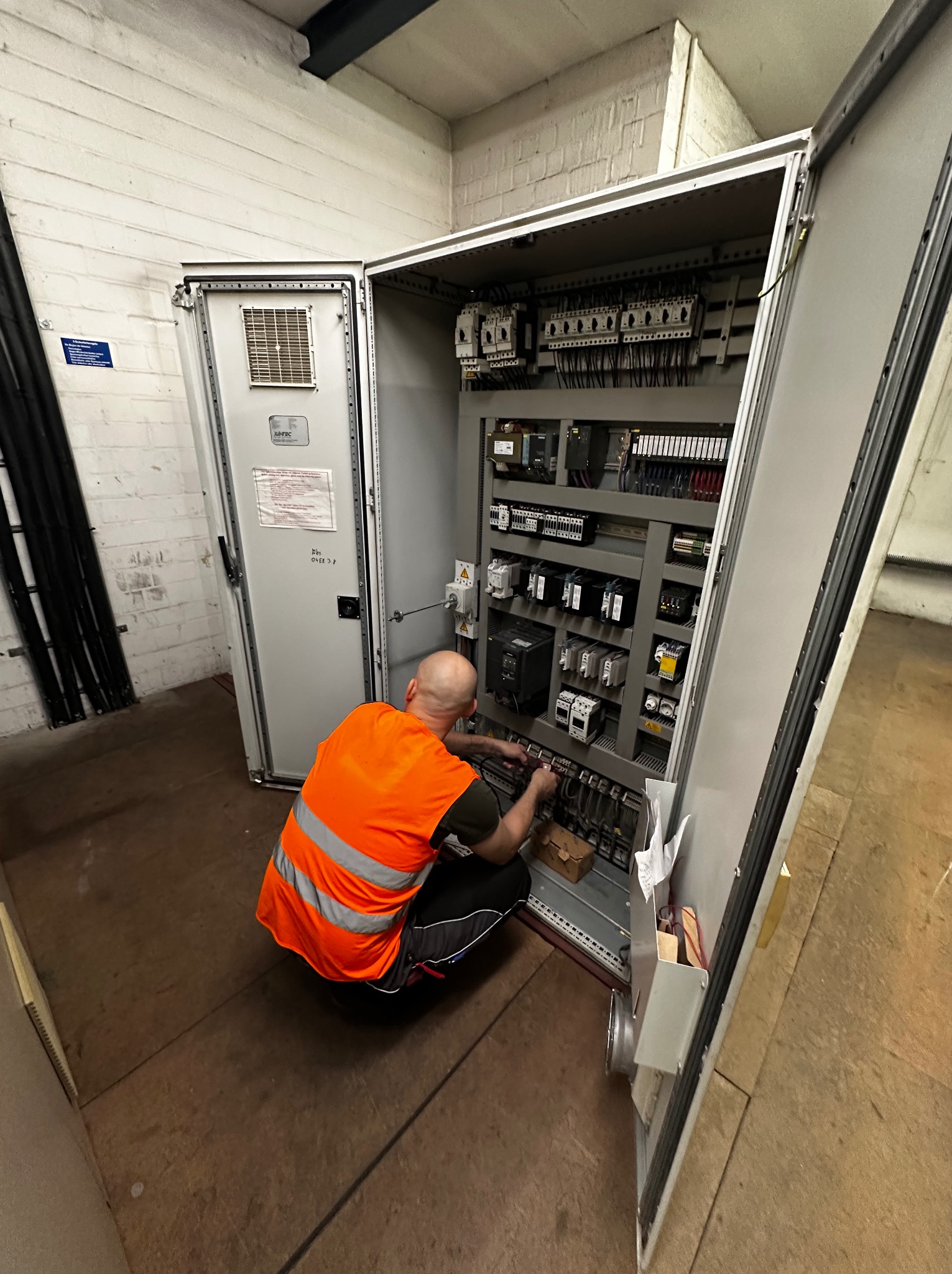 Elektryk w kamizelce BHP kuca przed otwartą szafą sterowniczą, wykonując prace instalacyjne. Widoczne liczne przewody i elementy elektryczne wewnątrz szafy.