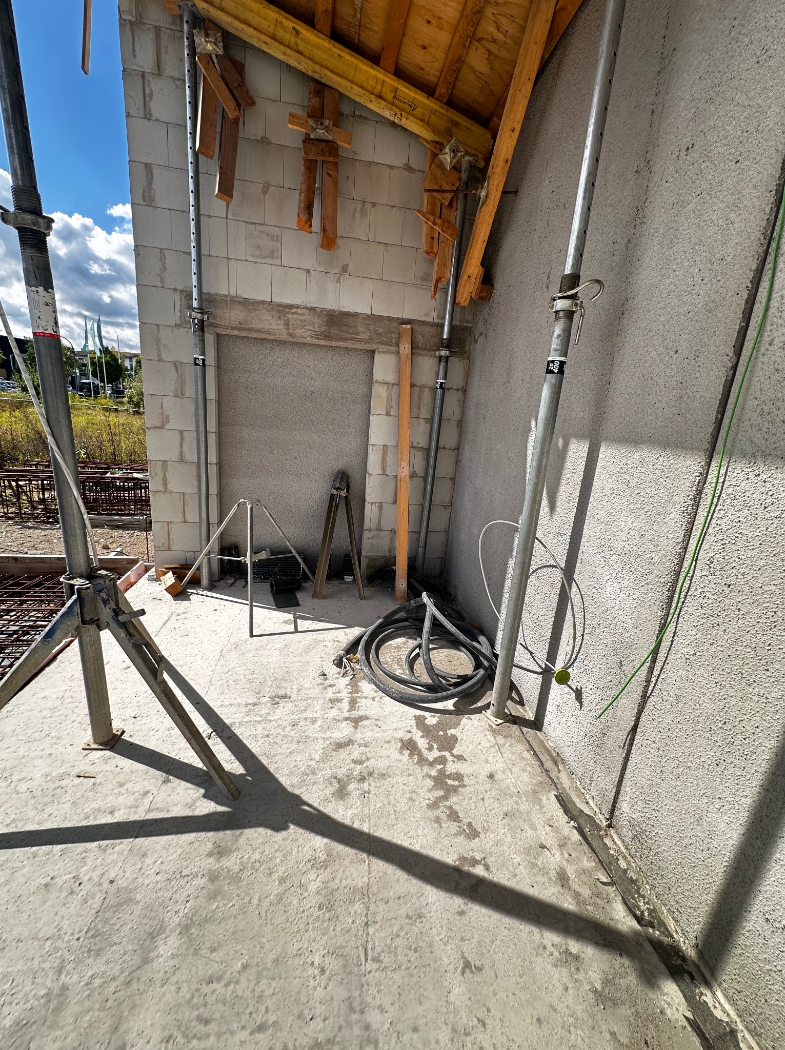 Wnętrze budynku w trakcie budowy: ściany z betonu komórkowego, drewniane stemple podtrzymujące strop, zwój kabli elektrycznych i narzędzia na betonowej posadzce.