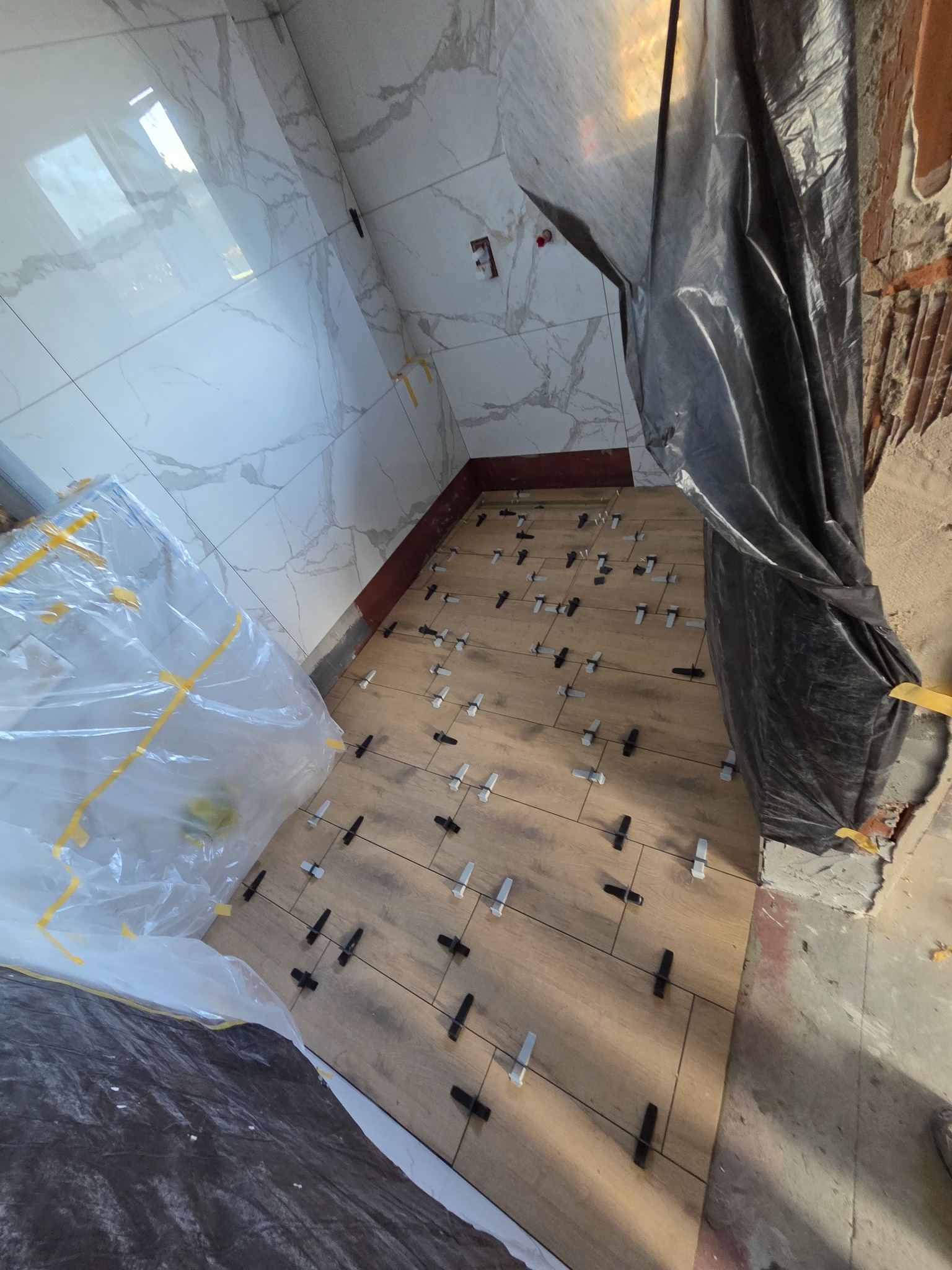 Układanie drewnopodobnych płytek podłogowych z użyciem klipsów poziomujących w łazience w Paszynie. Widoczne marmurowe płytki ścienne i zabezpieczenia folią.