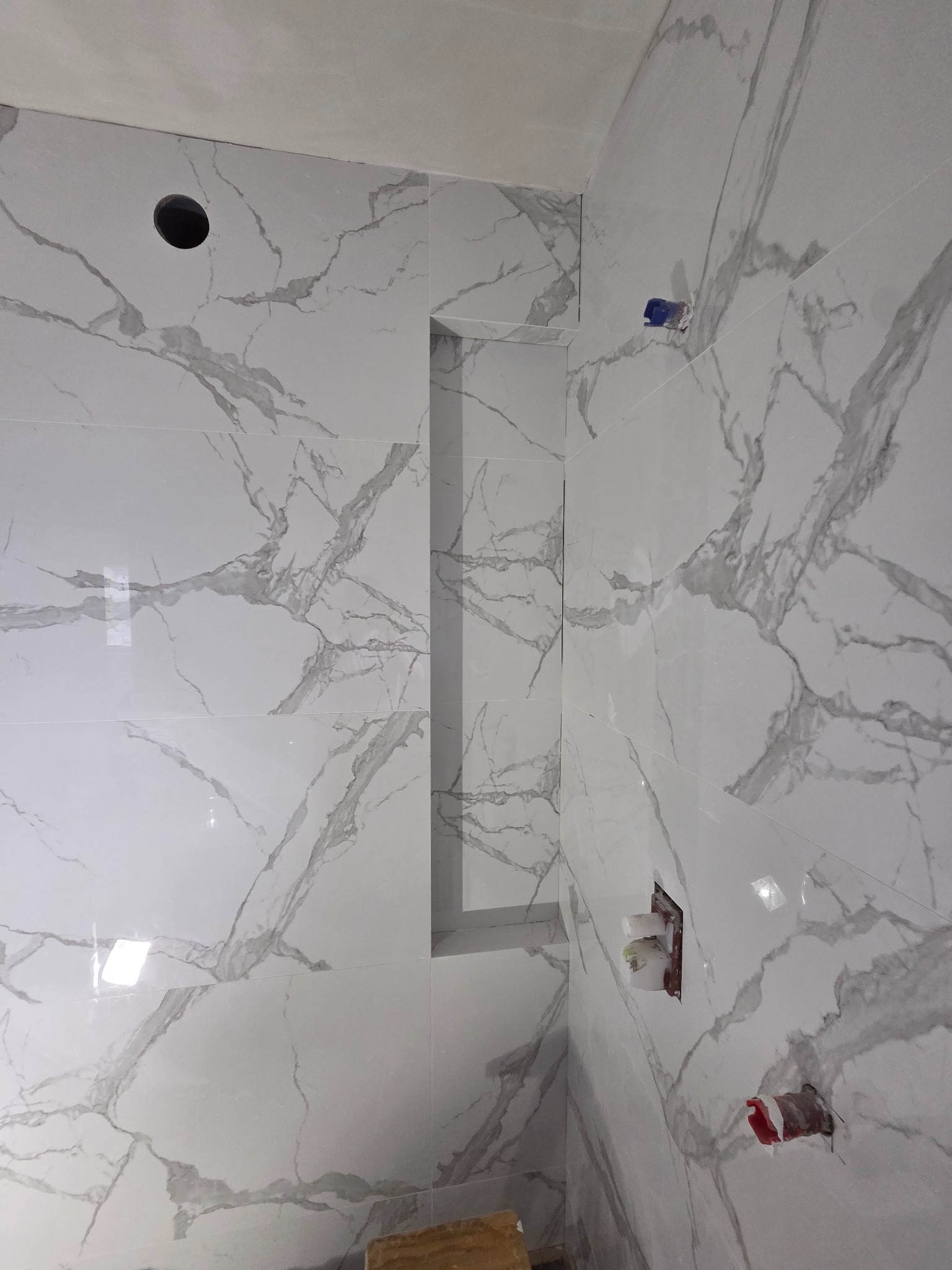 Wykończona ściana łazienki z białych płytek imitujących marmur, z wnęką i otworami na instalacje. Widoczne fragmenty instalacji wodnej i elektrycznej.