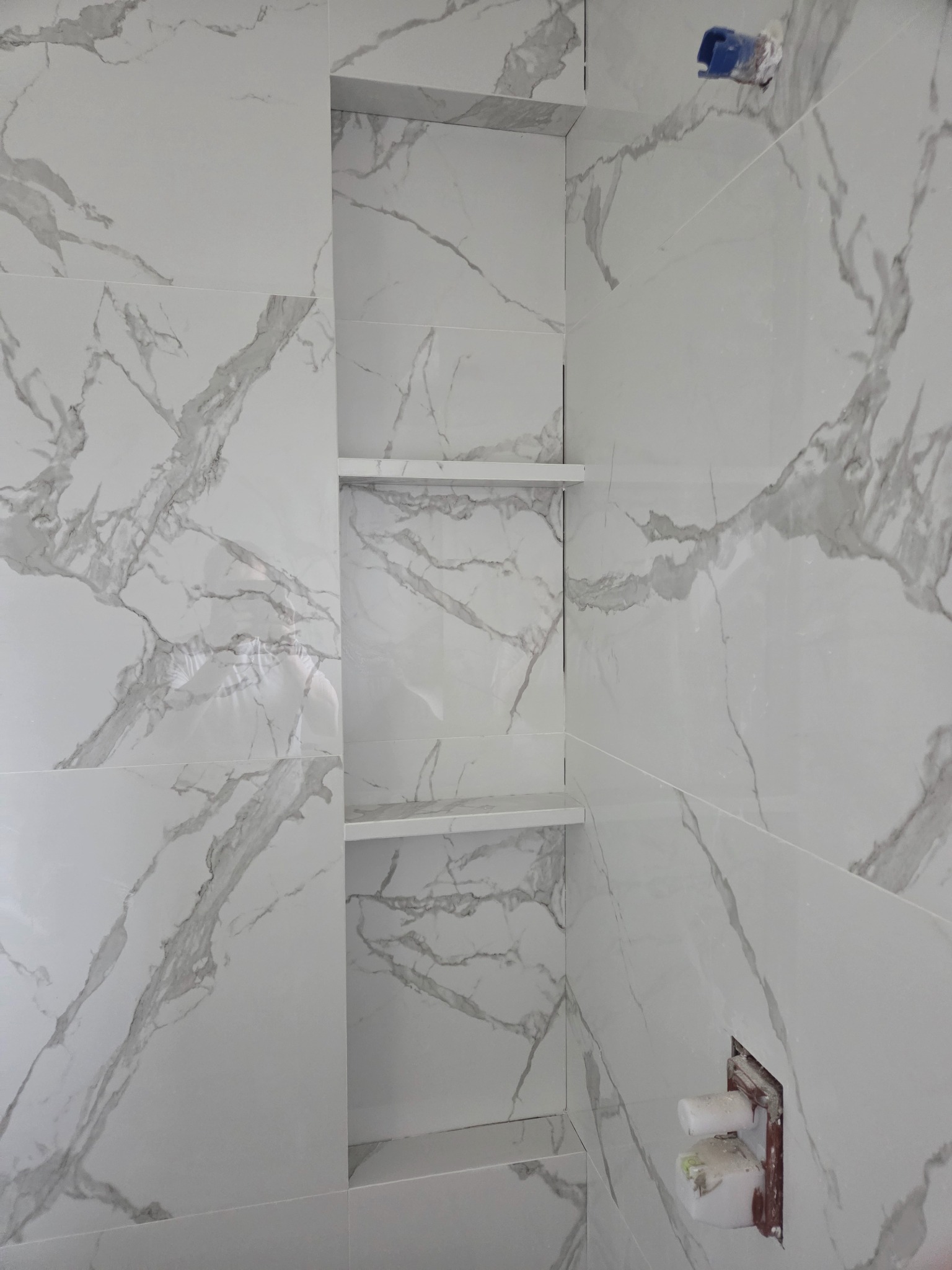 Biała łazienka z płytkami imitującymi marmur, wnęka z półkami. Widoczne elementy instalacji wodnej przed ostatecznym wykończeniem.