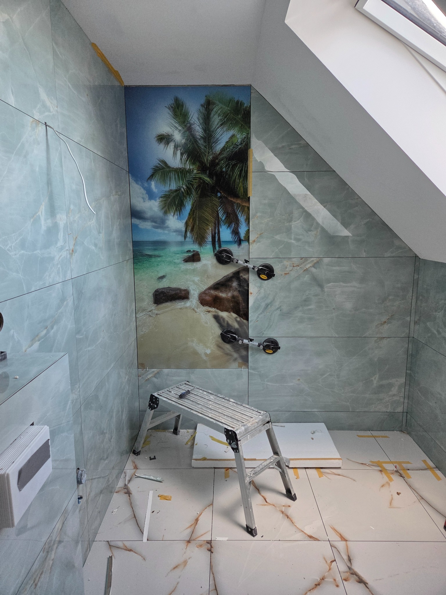 Układanie płytek w łazience na poddaszu. Na ścianie płytka z motywem plaży. Widoczna drabina i narzędzia glazurnicze. Praca w toku, wykończenie wnętrza.