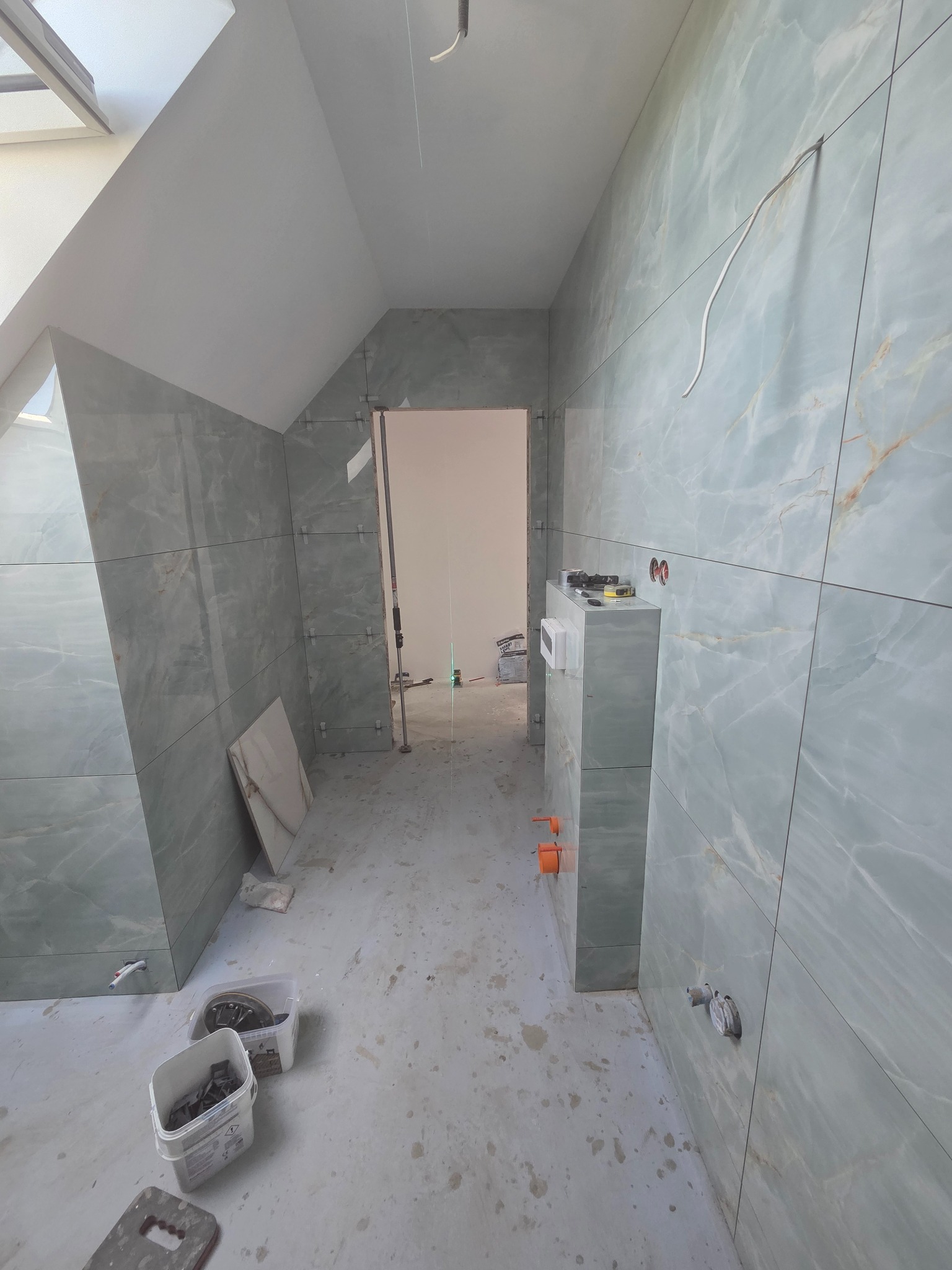 Łazienka na poddaszu w trakcie wykańczania: ściany wyłożone płytkami imitującymi marmur, widoczne narzędzia glazurnicze i instalacje. Skosy sufitowe.
