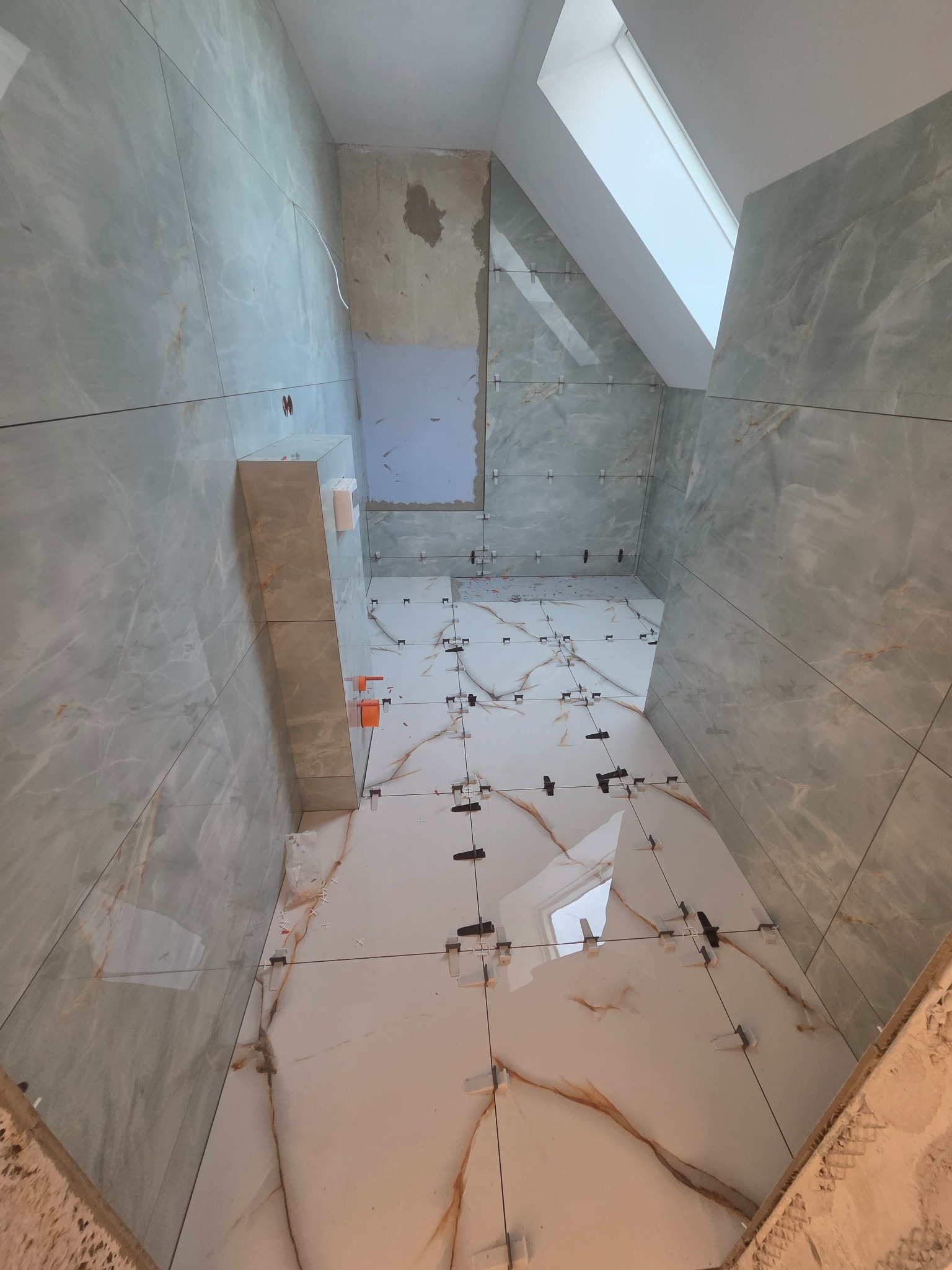 Układanie płytek w łazience na poddaszu z oknem dachowym. Widoczne klipsy poziomujące. Część ściany bez płytek. Płytki imitujące marmur w odcieniach szarości i bieli.