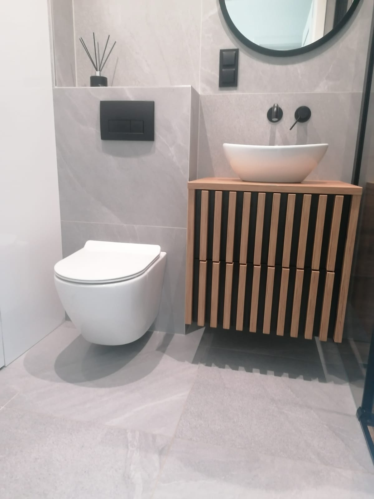 Nowoczesna łazienka z podwieszaną toaletą, umywalką nablatową na szafce z drewnianymi lamelami i szarą płytką imitującą marmur na ścianach i podłodze.