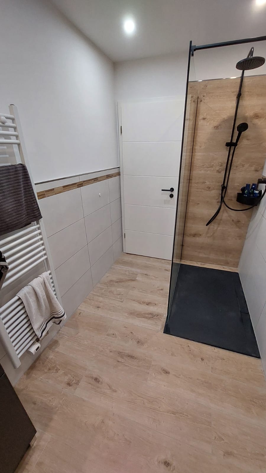 Nowoczesna łazienka z kabiną typu walk-in, drewnopodobną podłogą i grzejnikiem drabinkowym. Minimalistyczny design, białe ściany z akcentem dekoracyjnym.