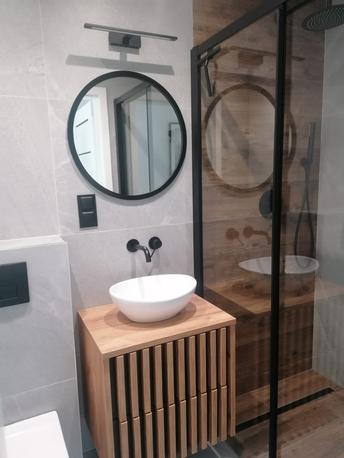 Nowoczesna łazienka z okrągłym lustrem, umywalką nablatową na drewnianej szafce i kabiną prysznicową z czarnymi profilami. Minimalistyczny design, szare płytki.
