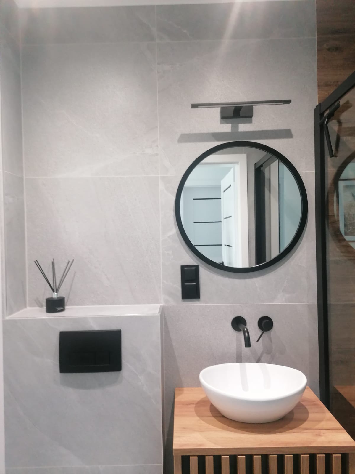 Wykończona łazienka z szarymi płytkami, okrągłym lustrem w czarnej ramie, drewnianym blatem i białą umywalką. Minimalistyczny design z czarnymi akcentami.