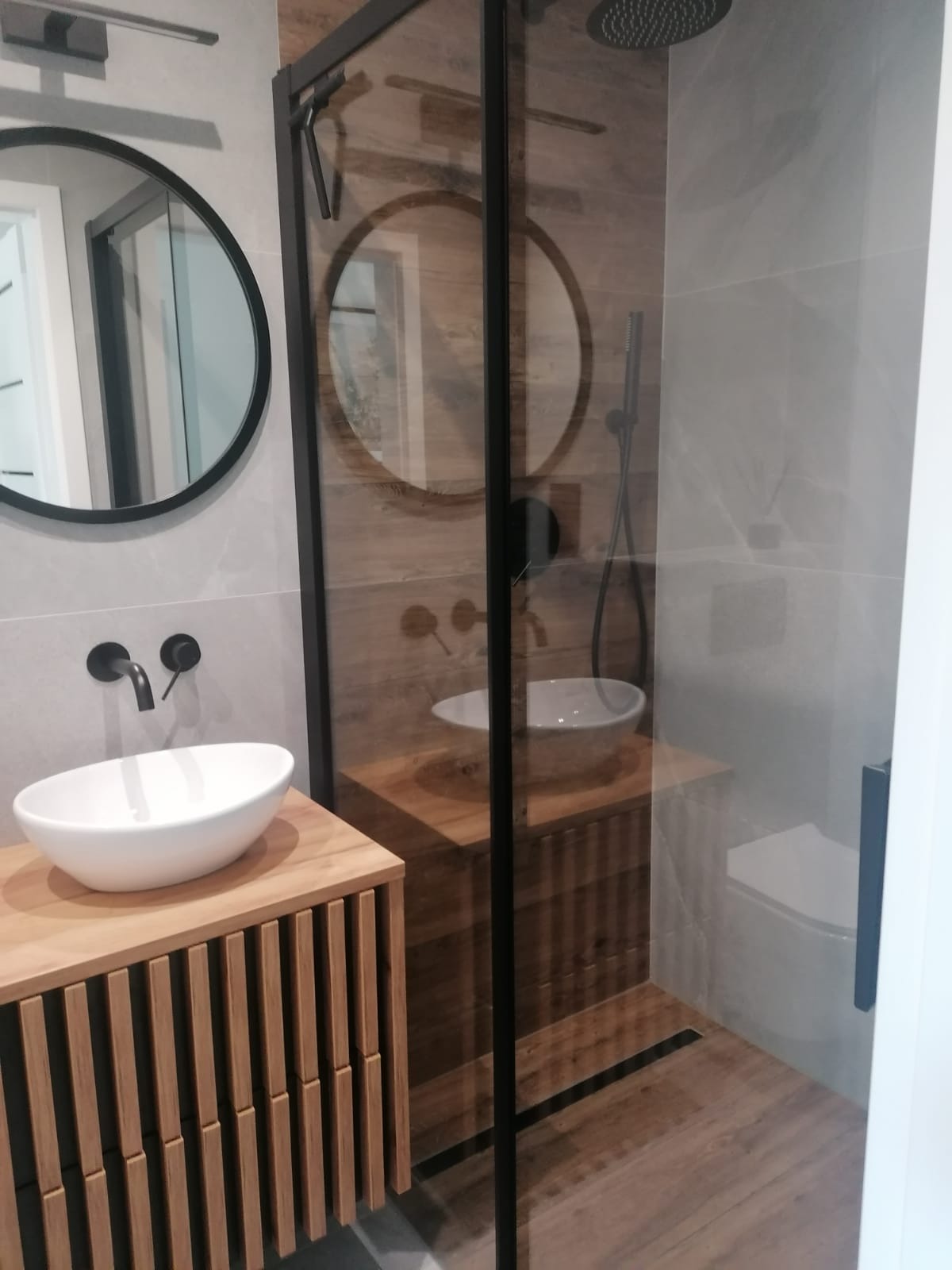 Nowoczesna łazienka z drewnianą szafką pod umywalką, okrągłym lustrem i kabiną prysznicową typu walk-in z czarnymi profilami. Minimalistyczny design.
