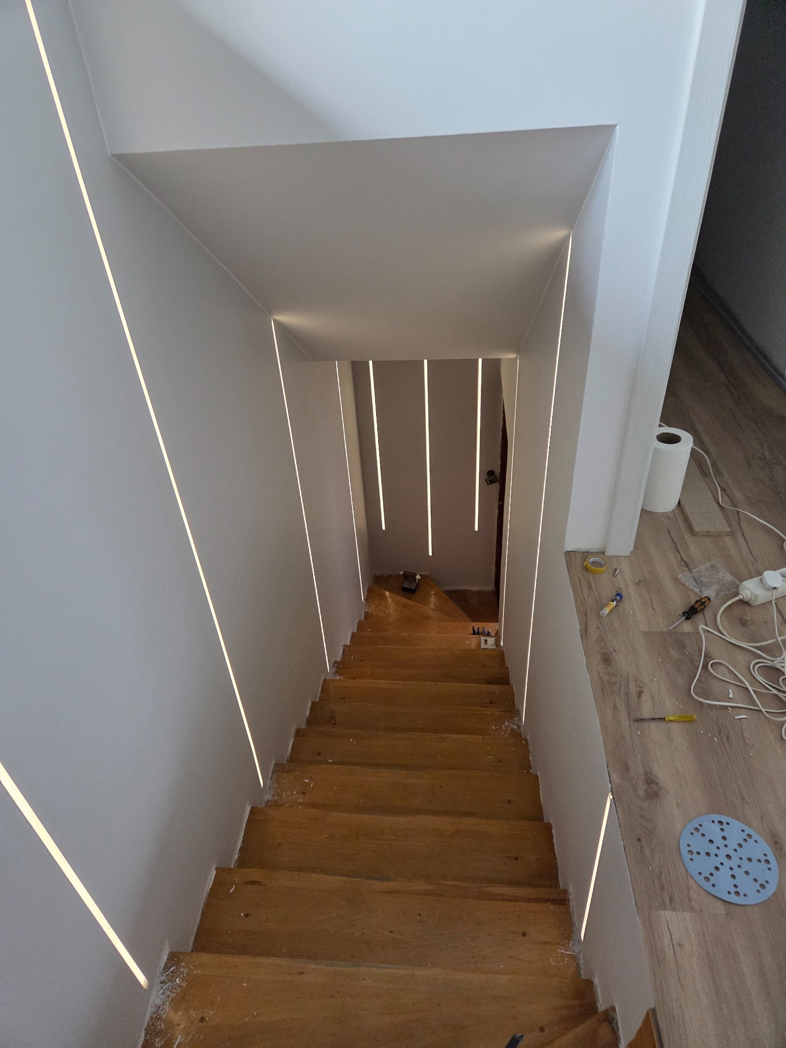 Schody drewniane z oświetleniem LED w ścianach, widok z góry. Widoczne narzędzia i materiały wykończeniowe, co sugeruje etap prac wykończeniowych.