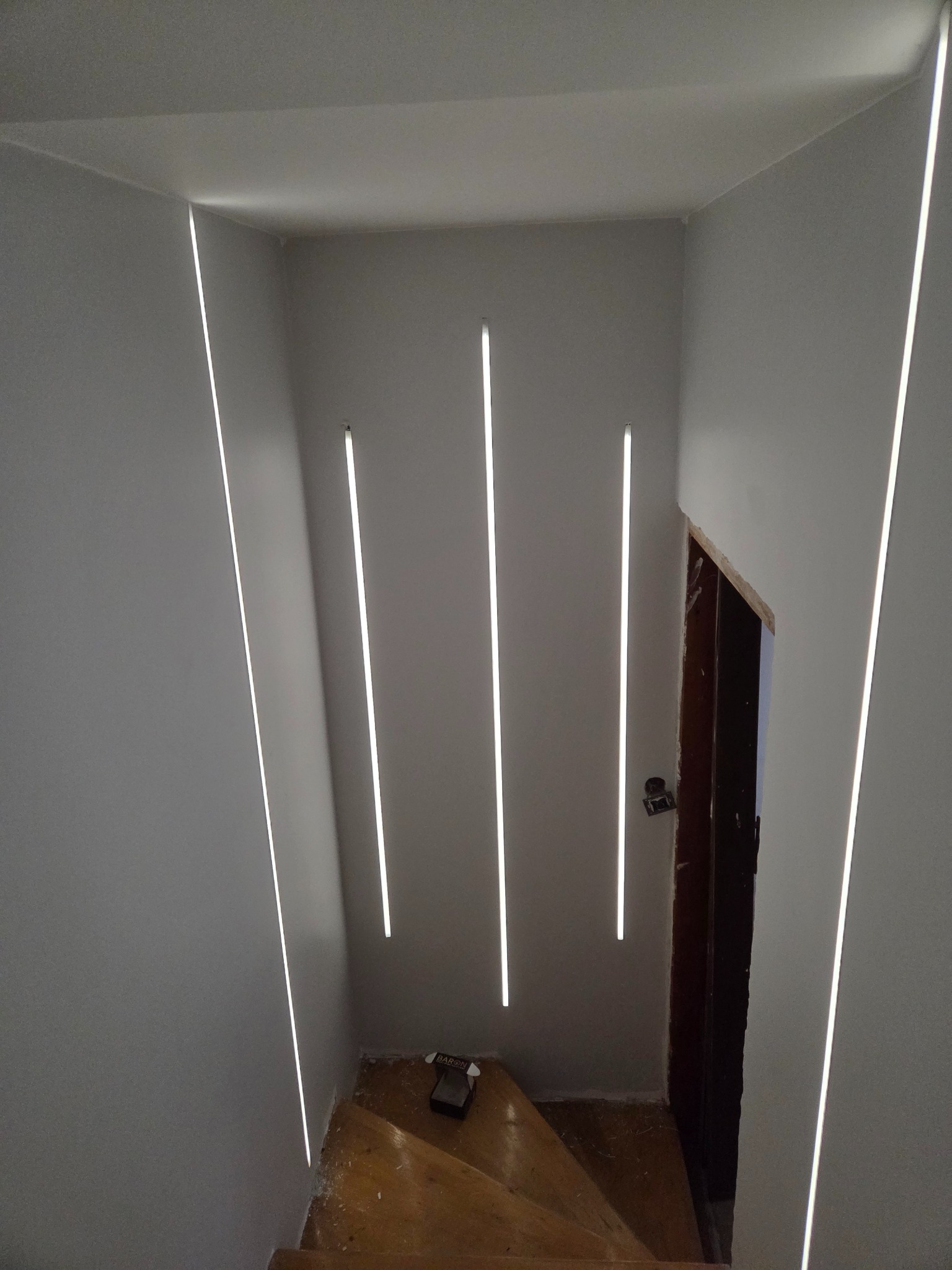 Nowoczesne oświetlenie schodów z wbudowanymi pionowymi lampami LED w ścianach. Widoczne drewniane schody i fragment otwartych drzwi.