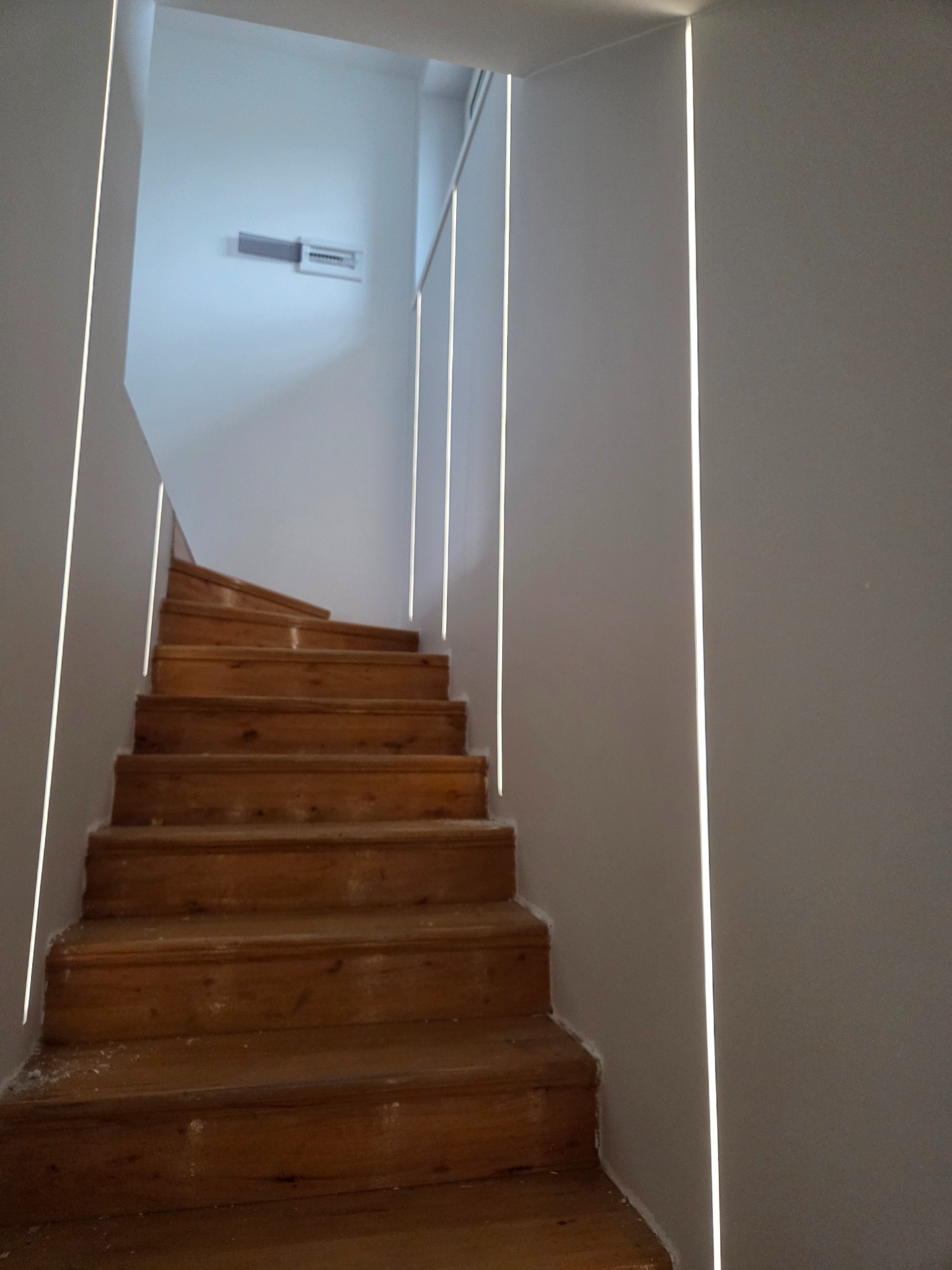 Drewniane schody z podświetleniem LED wzdłuż ścian, prowadzące w górę. Widoczne białe ściany i sufit. Ujęcie z dołu, podkreślające pionowe linie światła.