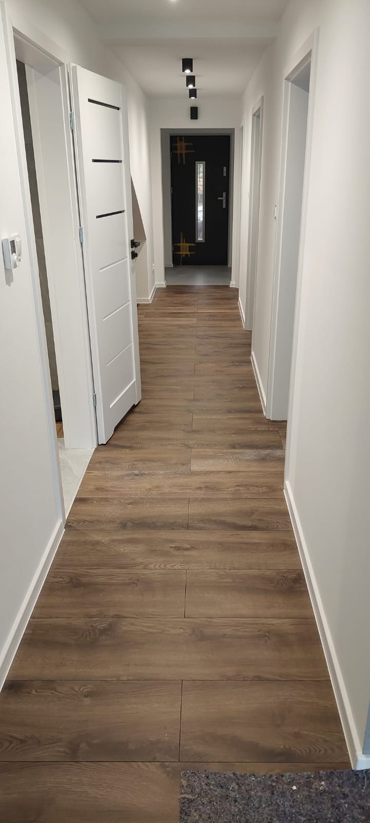 Korytarz z panelami podłogowymi imitującymi drewno, białe ściany i drzwi. Widoczny fragment podkładu pod panele w rogu kadru. Nowoczesne oświetlenie sufitowe.